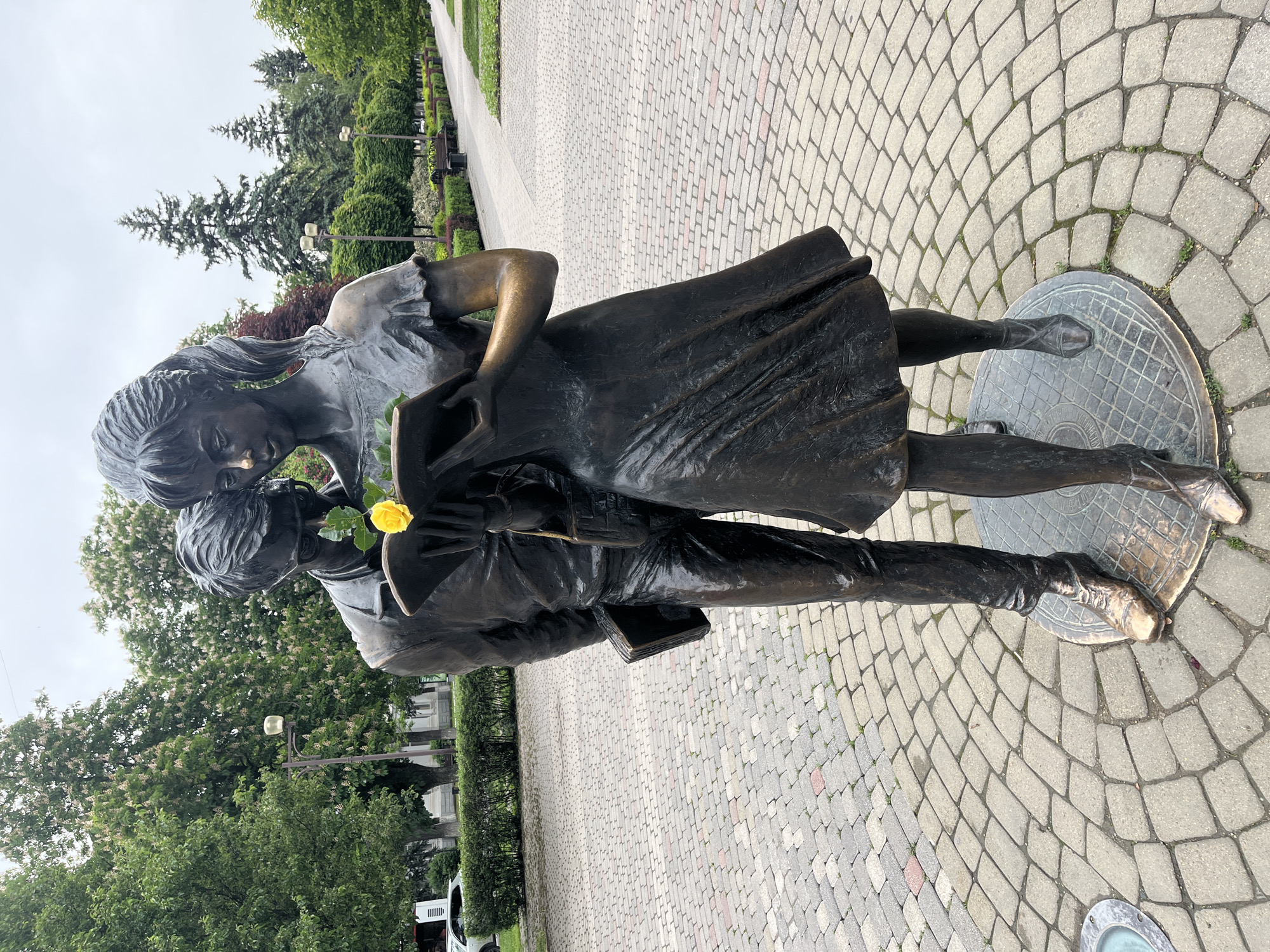 A bronze statue of a young couple stands on a circular metal base on a paved walkway. The statue depicts a girl in a dress leaning over a boy, possibly whispering to him. The boy holds a book, and a yellow flower is tucked near his head. The walkway is made of light-colored paving stones in a circular pattern around the statue. In the background, there is a park setting with trees, bushes, and benches. The weather appears overcast. The image is sideways, needing rotation for proper viewing. The statue is located in the city of Krasnodar, Russia.