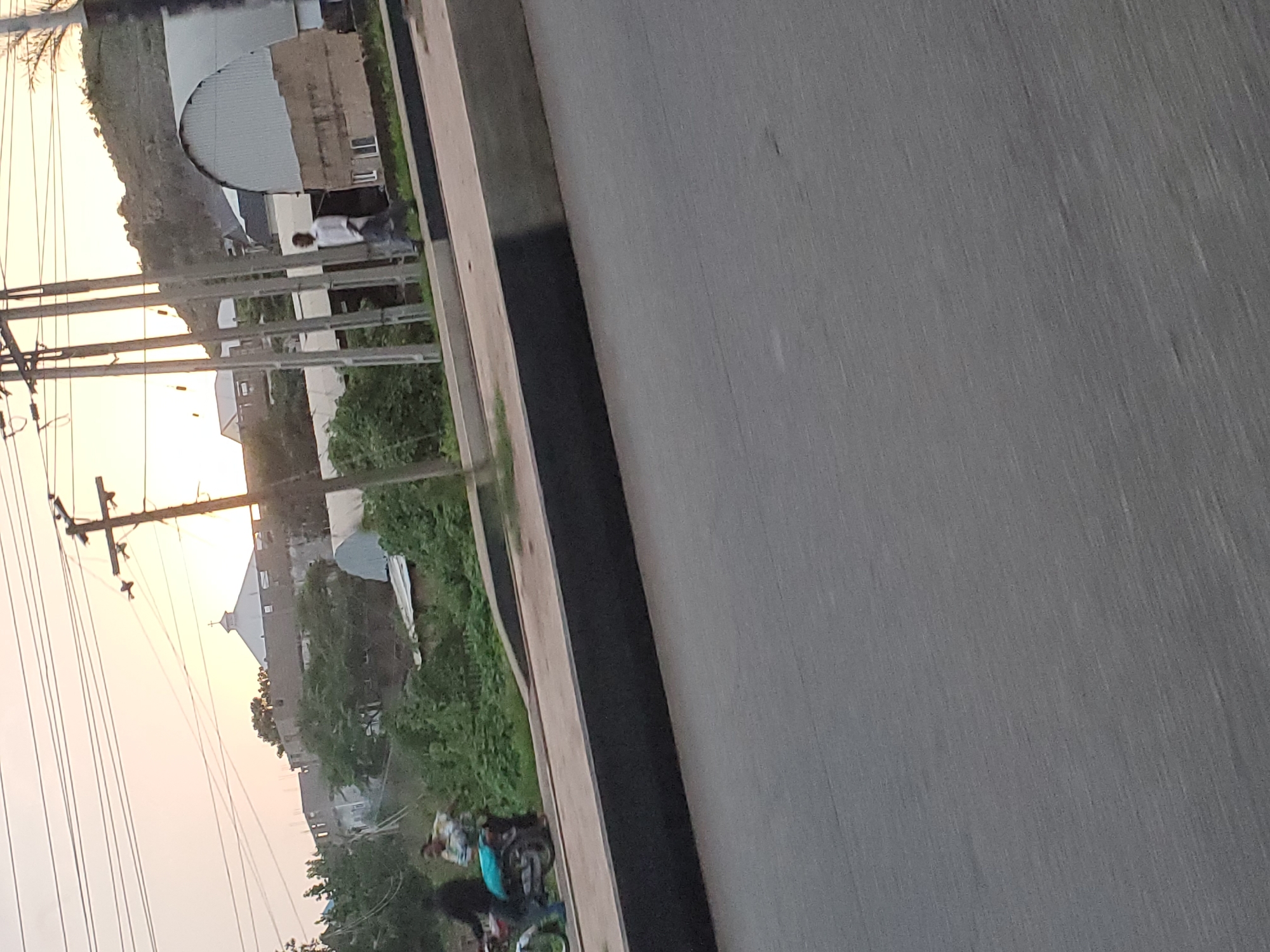 The image depicts a street scene at dusk in Gura Top, Nigeria. The sky is a soft orange and yellow, indicating sunset. In the background, a distinctive building with a white domed roof topped with a cross suggests a religious institution, possibly a church. Adjacent to it, a long, curved, grey-metallic structure with unfinished brickwork and small windows is visible. A lone man in a white shirt and dark pants walks on the sidewalk. To the left, a motorcycle with two people on it, one driving and another seated behind, is parked or stopped. The motorcycle has a bright blue bag strapped to its rear. The foreground is dominated by an asphalt road. A raised concrete walkway with a dark grey border separates the road from the area where the people and structures are located. Utility poles with multiple wires stretch across the sky. Lush green vegetation, including trees and bushes, borders the walkway.