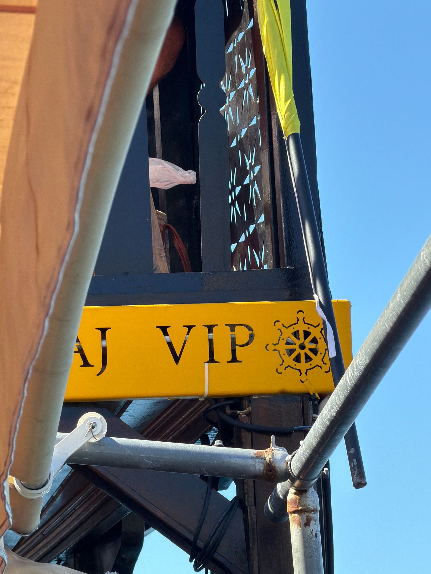 The image shows a close-up view of a yellow sign with black lettering that reads "AJ VIP". To the right of the text is a black, stylized ship's wheel emblem. The sign is mounted on dark wooden structures, possibly part of a boat or a waterfront establishment. There are also metal scaffolding pipes in the foreground and background, some of which appear rusty. A bright yellow fabric is draped over a dark pole to the right of the sign, against a clear blue sky. The overall scene suggests a location in Saranda, Albania, likely near a harbor or a venue that caters to VIP guests.