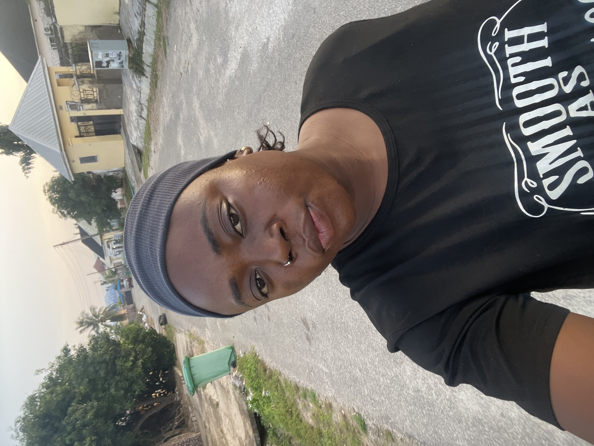 A woman in a black t-shirt and a grey beanie is taking a selfie in what appears to be a residential street in Abuja, Nigeria. The street is paved and appears to be at ground level. To the left of the woman, a green trash can is visible on the side of the street. In the background, there are several houses with yellow and white exteriors and corrugated metal roofs. Trees are also present on the left side of the image. The sky is overcast, suggesting it might be dawn or dusk. The woman has a nose ring and earrings. The t-shirt has white text that reads "SMOOTH AS" in a decorative font.