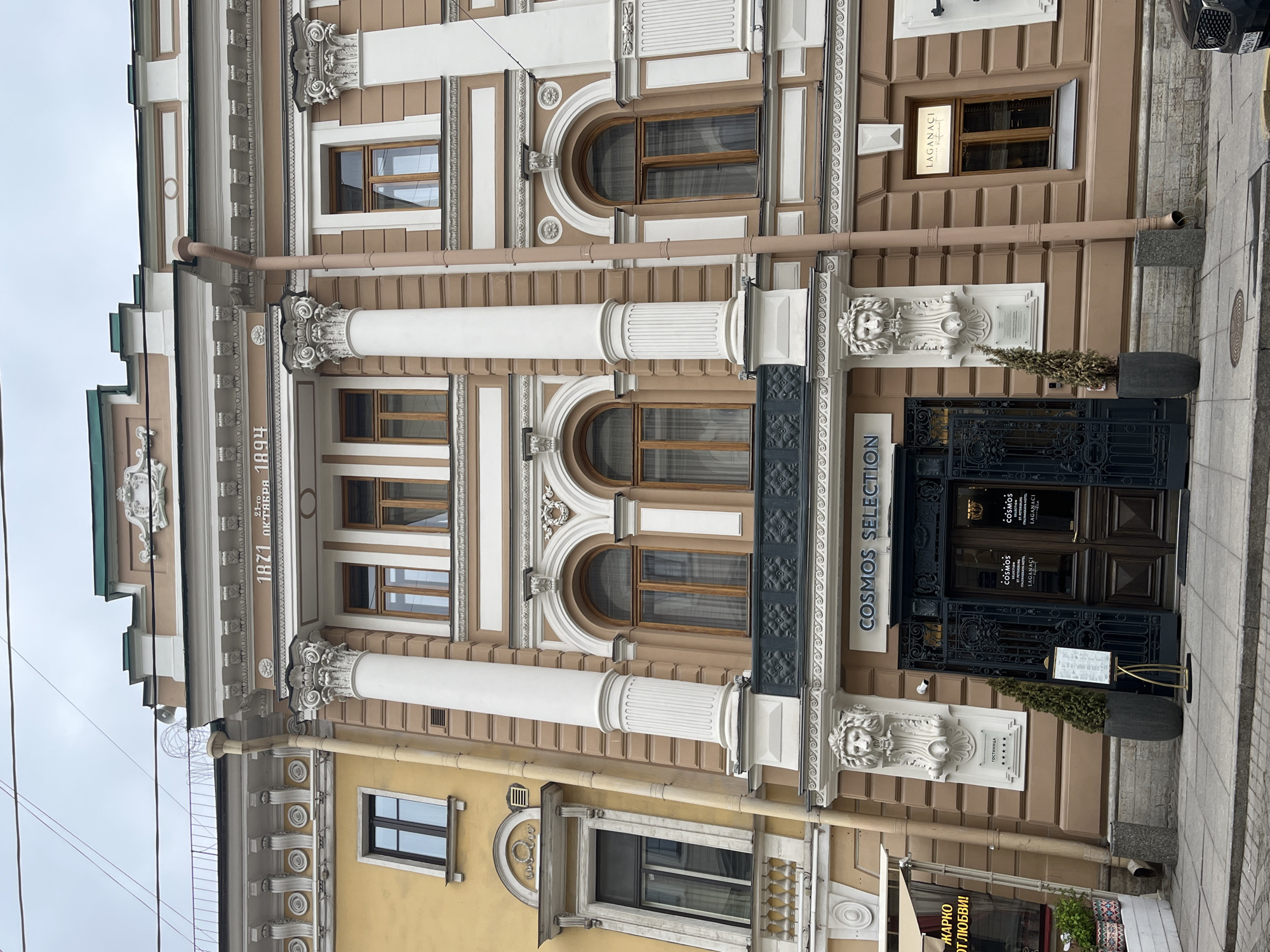 The image shows the facade of a building in Saint Petersburg, Russia. Architectural details are prominent, including decorative columns, arched windows, and ornate molding. The building appears to have a restaurant on the ground floor called "COSMOS SELECTION". The name "LAGANACI" is also visible on a sign. The numbers "1871" and "1894" are inscribed on the building's facade. The environment is an urban street, with parts of adjacent buildings visible on either side.