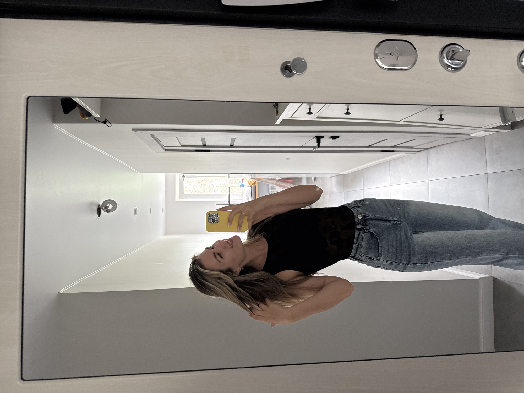 A blonde woman in a black top and blue jeans takes a mirror selfie in a hallway. She is smiling and holding her hair with one hand while holding her yellow phone in the other. The hallway has light gray walls and a tiled floor. A tall white cabinet with several drawers is to her right, and a door is further down the hallway. The mirror reflects the hallway and a window at the end.