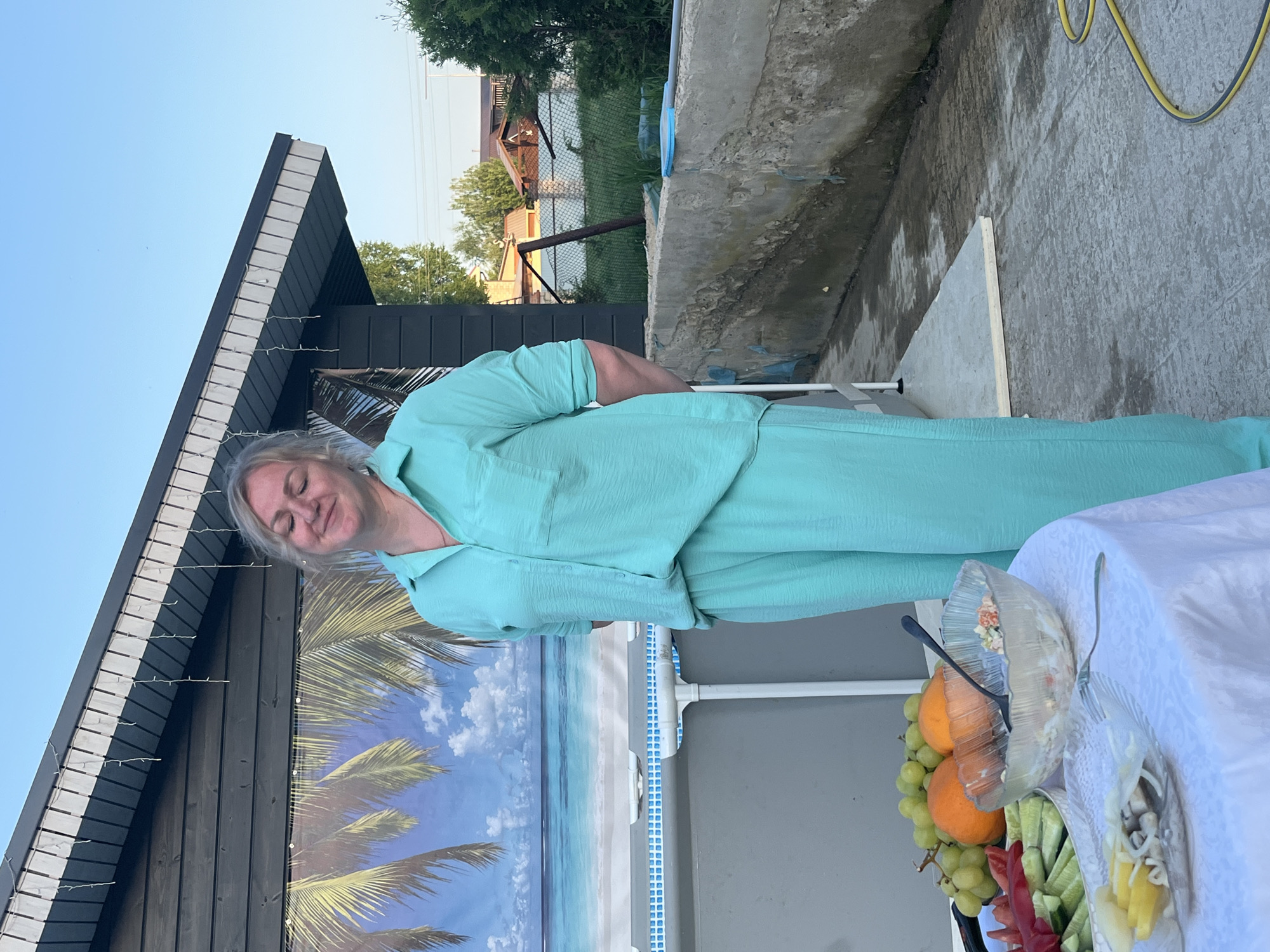 The image captures a woman in a light blue suit standing next to a table laden with food outdoors in Новая Чернушка, Russia. On the table, covered with a white tablecloth, are platters containing fruits, vegetables, and salads. Behind the woman, a dark wooden building features a palm tree backdrop and string lights along its roofline. A concrete structure and a wire fence with vegetation can be seen in the background. The sky is clear and blue.