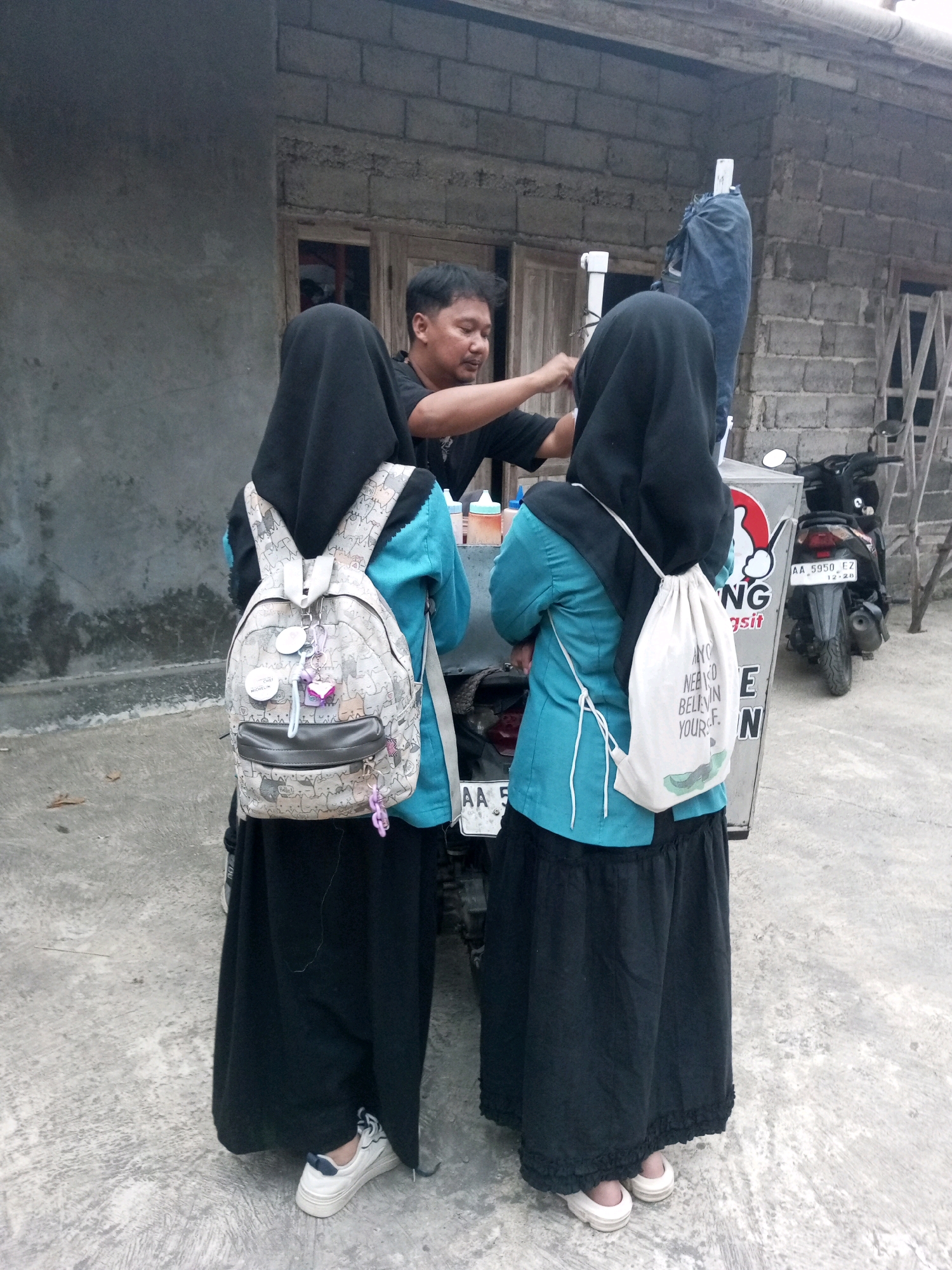 In Krasak, Indonesia, a man wearing a black t-shirt is interacting with two young women wearing hijabs and long black skirts, with one woman wearing a turquoise top and the other a black top. The man appears to be attending to something near a vendor cart. Both women have backpacks on. The woman in the turquoise top has a light-colored drawstring bag hanging from her shoulder, and the woman in the black top has a patterned backpack with several charms. A motorcycle is parked behind the vendor cart. The environment suggests an outdoor, possibly commercial, setting with a brick building in the background.