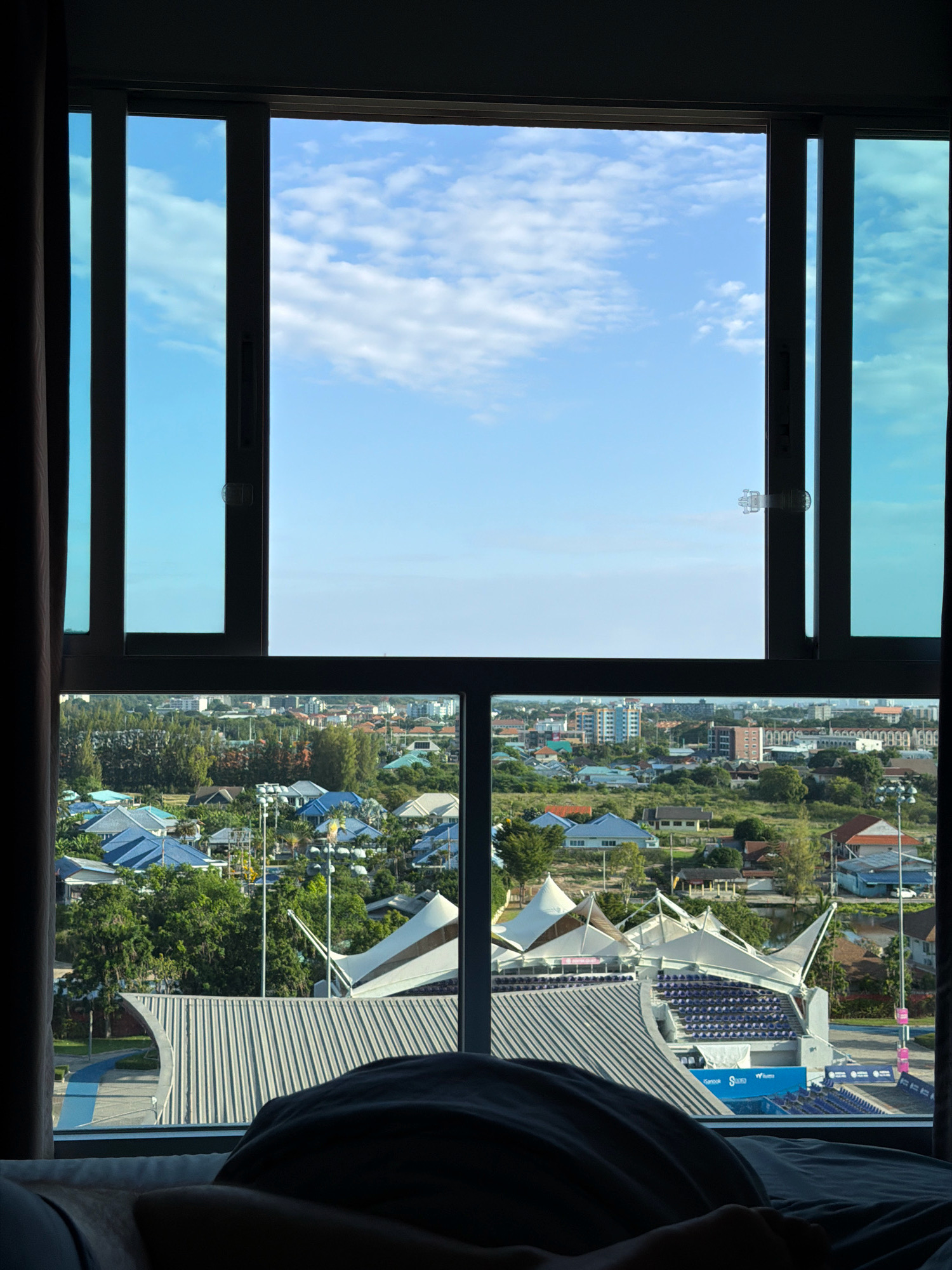 The image, taken from a hotel room in Hua Hin City Municipality, Thailand, looks out through a large window onto a cityscape and a bright blue sky with scattered white clouds. The window is divided into two sections. The upper section, which is open, reveals a clear blue sky with a few wispy clouds. The lower section of the window shows a dense residential area with numerous buildings, many with blue tile roofs, interspersed with lush green trees. In the foreground of the lower section, a large, modern stadium structure with white, tent-like roofs and rows of blue seating is visible. Part of a bed with dark grey bedding is also visible in the bottom portion of the image, suggesting the photograph was taken from someone lying in bed. The overall scene depicts a sunny day in a coastal city with a mix of residential housing and modern recreational facilities.