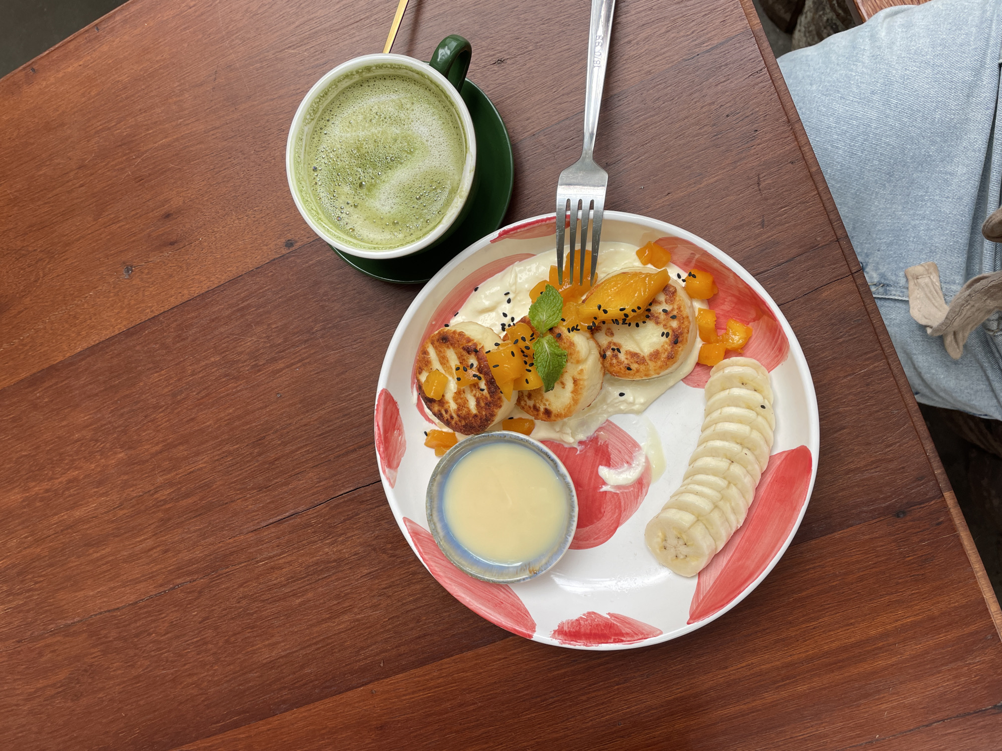 A close-up, top-down shot shows a plate of syrniki, a type of Russian cheese pancake, alongside a cup of matcha latte, both resting on a wooden table. The syrniki are arranged in a stack of three, topped with diced mango, a sprig of mint, and sprinkled with black sesame seeds. A small dish of condensed milk sits to the left of the pancakes, and sliced bananas are positioned at the bottom left of the plate. The plate itself features a red, abstract brushstroke design. The matcha latte is a vibrant green with a frothy top and is served in a white cup with a green saucer. A fork rests on the edge of the plate, angled towards the syrniki. The wooden table has a visible grain and a few subtle cracks. The image is taken from a high angle, with the bottom right corner of the frame showing a person's light blue denim jeans and a beige leg warmer. The scene appears to be set in a cafe or restaurant.