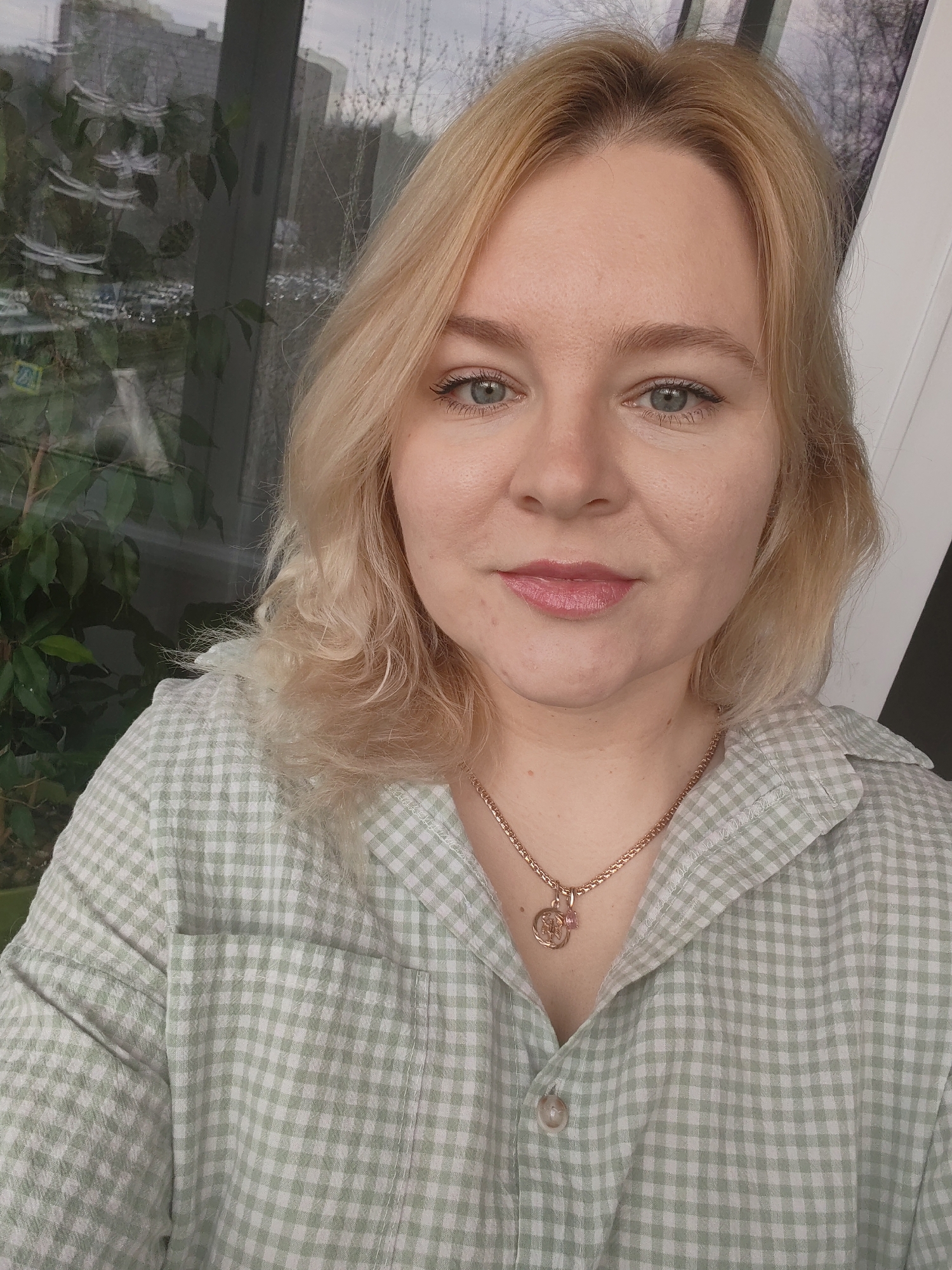 A woman with blonde, shoulder-length hair, blue eyes, and fair skin is centered in the frame. She is wearing a light green and white checkered button-down shirt and a gold necklace with a pendant. The background shows an indoor space with a window. The window looks out onto a cityscape, where some buildings and bare trees can be seen. There are also green plants visible near the window inside the room. The woman is facing the camera, and her expression is neutral. Given the available information, there is nothing in the image to indicate the location is in Izhevsk, Russia.