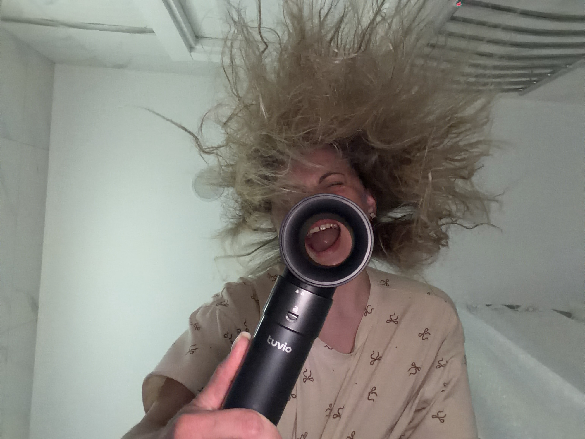 An indoor, close-up, and humorous shot features a person with wildly disheveled, blonde hair that stands out in all directions, creating an almost static electricity effect. The person's face is largely obscured by a black handheld device, held by their right hand which extends into the frame from the lower right.
The black device, branded "tuvio" in white text on its side, is cylindrical with a circular opening at the top. The person's wide-open mouth, revealing teeth and the tongue, is perfectly framed within this circular opening, conveying an expression of laughter, surprise, or a playful scream. The person's eyes are visible just above the device, partially closed as if in mirth.
They are wearing a light-colored garment, possibly a robe or pajamas, adorned with a repeating pattern of small, dark brown bow-like shapes.
The background consists of light-colored walls, possibly white or off-white, suggesting an interior space like a bathroom or a brightly lit room. A metallic grate or vent is visible in the upper right corner, further hinting at a utility area. The lighting is bright and even. The activity captured appears to be a playful moment, possibly involving a hairdryer or another styling tool that has left the hair in such a state, or it's a deliberate comedic pose. The image was captured in Votkinsk, Russia.