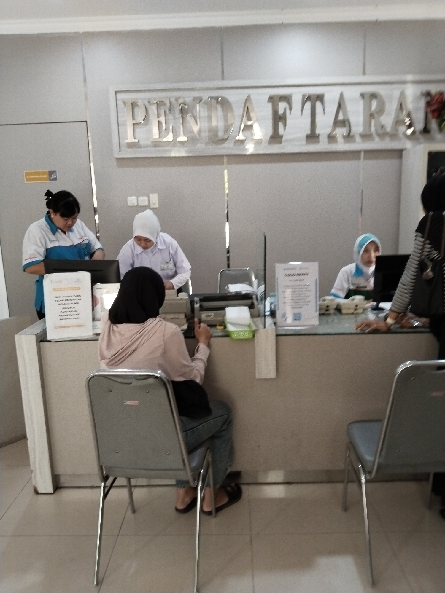 The image captures an indoor scene at a registration counter, likely a hospital or clinic, in Kecamatan Pati, Indonesia. A sign above the counter reads "PENDAFTARAN," meaning "REGISTRATION." Three women are visible. Two are employees behind the counter, and one is a customer sitting in a chair facing away from the camera.
The employee on the left, wearing a blue and white uniform, is looking down at a computer monitor. The employee in the middle, wearing a white hijab and a light purple uniform, is also looking down, seemingly at papers or a computer. The customer, wearing a light pink hoodie and jeans, is seated and has her back to the camera. She appears to be interacting with someone behind the counter.
The counter area is equipped with a computer, a printer, a scanner, and office supplies. The overall environment appears to be a functional, sterile, and well-lit registration area. The flooring is tiled. The lighting is overhead, creating a somewhat flat illumination across the scene.