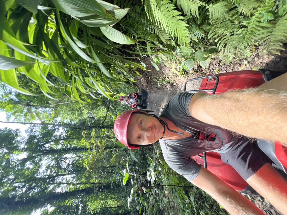 A fair-skinned man wearing a pink helmet and grey t-shirt is on a red ATV. The man's legs are tan and hairy, and sunglasses hang on his shirt. Behind him, another ATV is visible on the dirt path surrounded by dense green vegetation, including ferns and large-leafed plants. The lighting suggests a sunny day. The photo is taken from a first-person perspective while slightly tilted to the left.