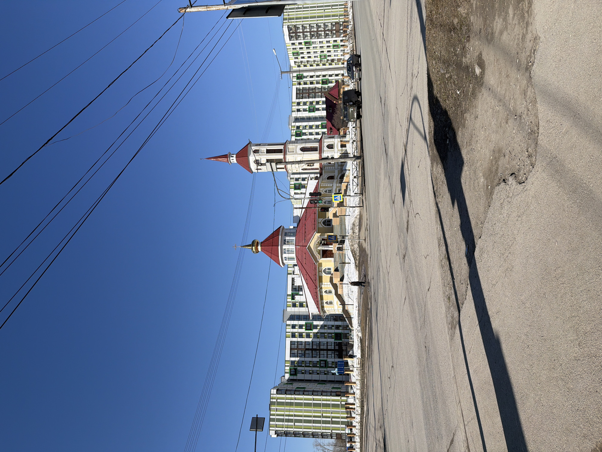 A low-angle, sunlit outdoor shot depicts a street scene in Berezniki, Russia, featuring a distinct church architecture set against a backdrop of modern residential buildings. The church, with its red-roofed domes and white-and-yellow facade, stands adjacent to multi-story apartments characterized by their white and green exteriors.
A paved road stretches along the right side of the frame, exhibiting patches of asphalt and casting long shadows from streetlights. Utility poles and power lines intersect the blue sky above. A single person is visible walking along the sidewalk near the church. Two military looking vehicles are parked in front of the church. The location appears to be a blend of urban development and traditional religious architecture.