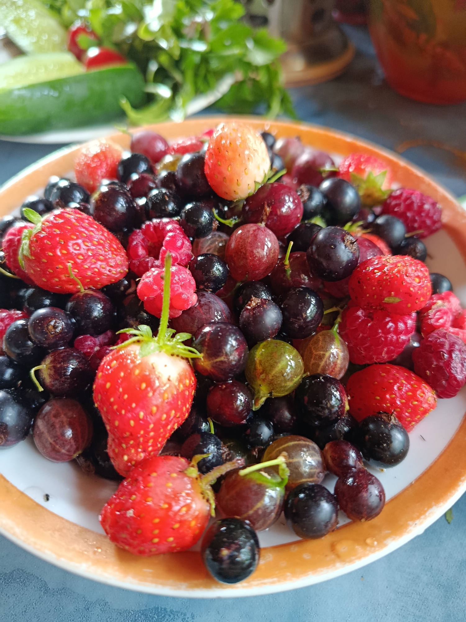 A close-up, high-angle shot shows a white plate filled with a variety of fresh berries, including strawberries, raspberries, blueberries, and gooseberries. The berries are glistening with water droplets, suggesting they are freshly washed. The plate has an orange rim. In the blurred background, a slice of cucumber and some greenery can be seen. The scene is lit from above.