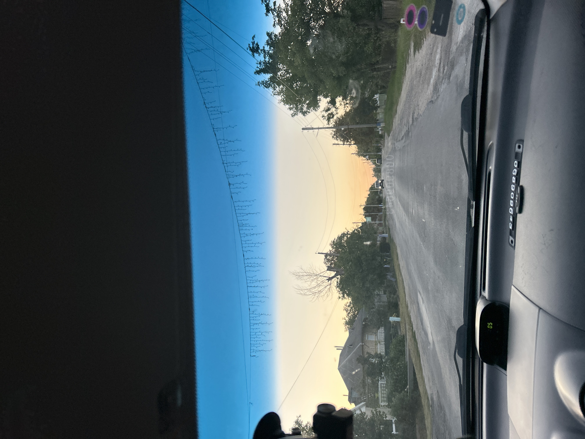 The image depicts a view from inside a vehicle driving down a paved road in Abaclia, Moldova, during what appears to be late afternoon or early evening due to the warm, soft light and the color of the sky. The road is lined with houses and trees on both sides.
In the foreground, the dashboard of the car is visible. A digital display shows "05". The windshield shows a string of lights hanging across the upper portion, possibly decorative.
The road itself is gray and appears to be in moderate condition, with some patches and wear. On the left side of the road, there are houses with pitched roofs and fences. On the right side, there are more trees and some wooden fencing.
In the middle distance, several cars are visible on the road, moving in the same direction as the viewer. The sky transitions from a bright blue at the top to a soft orange and yellow gradient near the horizon, indicating sunset or sunrise. Power lines and poles are present along the road.
The environment suggests a rural or semi-rural setting within the town of Abaclia. There are no people clearly visible in the scene. The overall atmosphere is calm and peaceful, characteristic of a drive through a residential area as the day winds down.