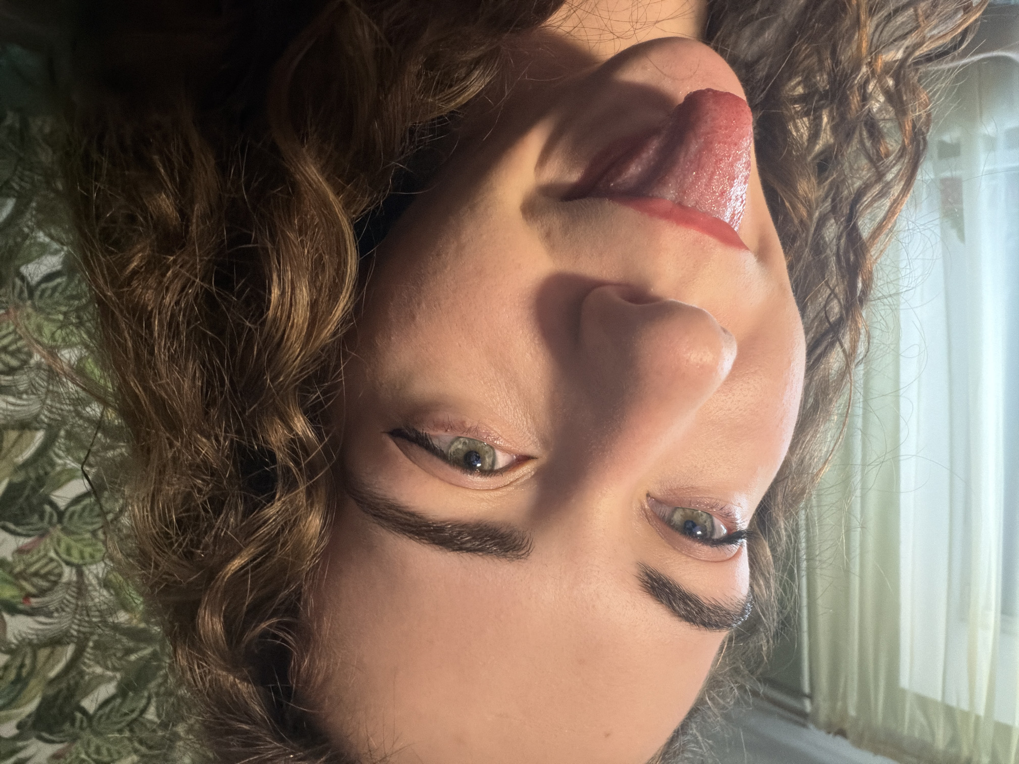 A close-up, eye-level, medium shot shows a young person with curly brown hair and green eyes. They are looking directly at the camera, with their tongue sticking out and colored red with lipstick. The background consists of a sheer curtain on the left and a green floral wallpaper on the right. The image is lit with warm, directional lighting, creating highlights on the person's skin and hair. The location is identified as Zavodoukovsk, Russia.