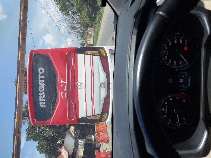 A forward-facing, eye-level photograph captures the rear of a red and white passenger bus on a paved road in Kulu, Indonesia, viewed from the interior of another vehicle. In the foreground, the upper portion of a black steering wheel and the vehicle's dashboard are visible. The instrument cluster displays a speedometer at 0 km/h, a tachometer at 0 RPM, the gear selector in Neutral ('N'), and an odometer reading 095918 km, with a green indicator light illuminated.
The bus, positioned directly ahead, features "ARIGATO" in white capital letters across its top rear window and "CJT" below it. Its lower rear section is white with red horizontal stripes, centered by a silver Mercedes-Benz logo. Yellow tail lights are visible at the bottom corners.
To the left of the bus, orange traffic barriers line the roadside, behind which a building with a reddish-brown roof and green foliage is visible. An overhead structure, appearing to be a wooden or metal beam, spans horizontally across the upper frame, above the bus. The road extends into the background on the right, bordered by trees, utility poles with power lines, and distant buildings, all under a clear, bright blue sky with scattered white clouds. The scene suggests a stationary position for the viewer's vehicle behind the bus during daylight hours.