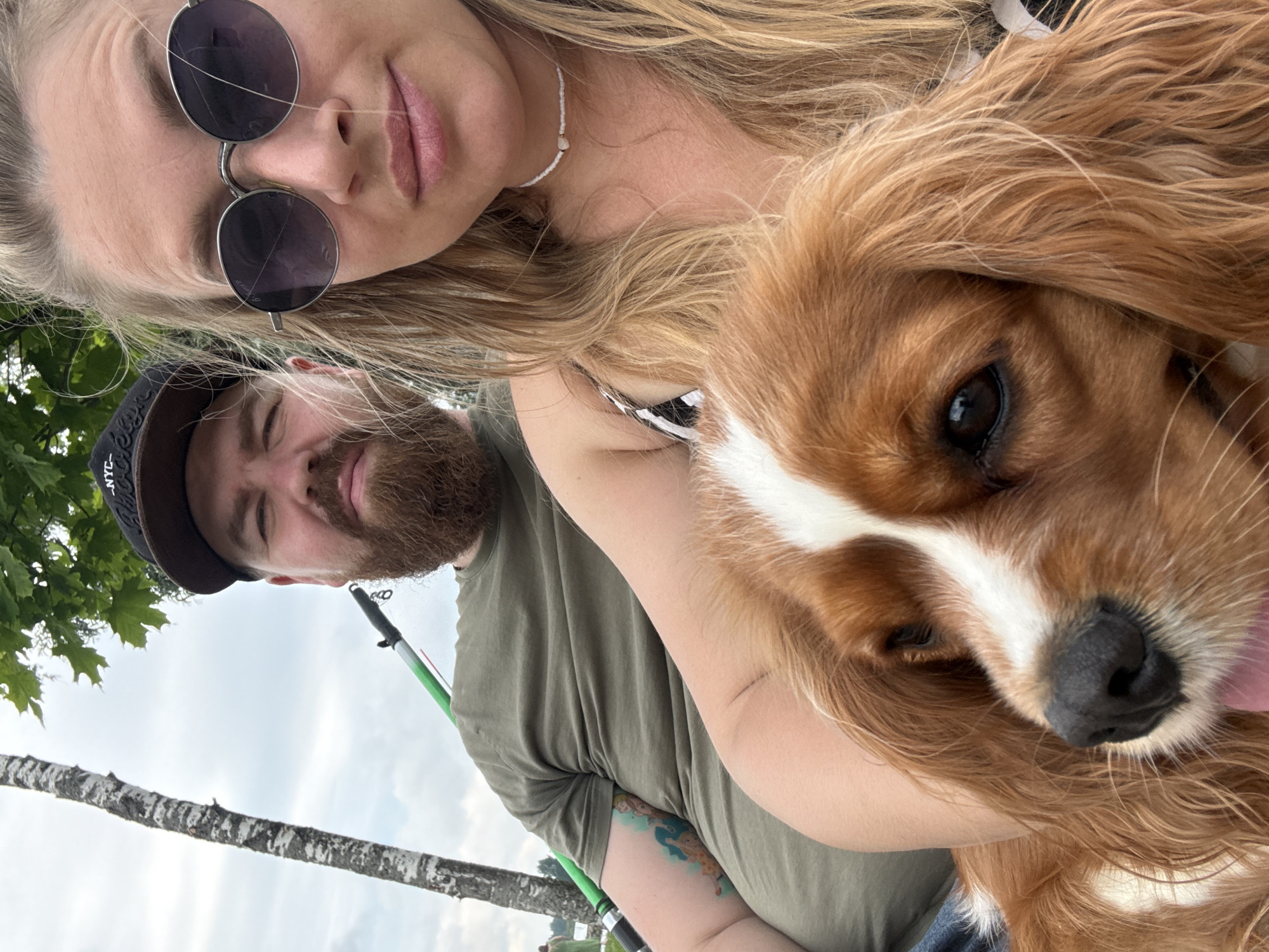 A group selfie shows a man and a woman posing with a dog. The man, wearing a cap and a green t-shirt, has a beard and a tattoo on his arm. The woman, wearing sunglasses and a patterned top, has blonde hair and is wearing a beaded necklace. The dog, a Cavalier King Charles Spaniel, is held by the woman. In the background, a tree and foliage are visible, suggesting an outdoor setting. The overall scene appears to be a casual outdoor gathering. It is not possible to determine the exact location within Valday, Russia from the provided image.