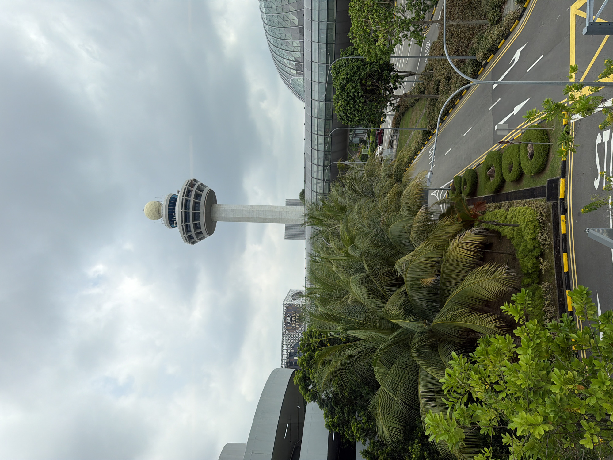 The image shows a view of Singapore Changi Airport, specifically the air traffic control tower and the Jewel Changi Airport complex, under a cloudy sky. The air traffic control tower, a tall, slender structure with a distinctive spherical top, stands prominently in the background. To its right, the large, domed structure of Jewel Changi Airport, with its glass facade, is visible. In the mid-ground, a multi-level car park with a curved facade is on the left, and a covered walkway or bridge connects different parts of the airport. Lush green palm trees and other tropical vegetation are abundant in the foreground and mid-ground, bordering a paved road with clear lane markings and yellow and black safety barriers. The road appears to be an airport access road or a service road. There are no people visible in the frame. The overall impression is one of a modern, well-maintained airport environment with a significant amount of greenery.