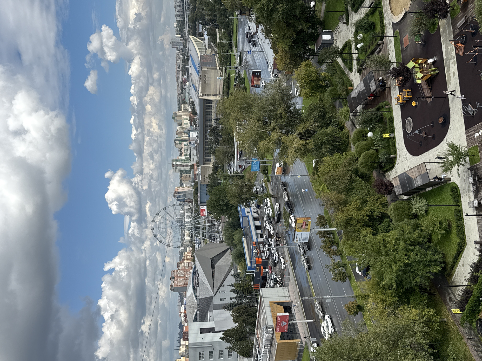 The image captures a panoramic view of an urban landscape in Chelyabinsk, Russia. In the foreground, a modern playground with various play structures, including slides, swings, and climbing equipment, is situated on dark rubberized flooring. Adjacent to the playground is a sandpit and an area with outdoor fitness equipment. Beyond the playground, a paved walkway is lined with lush green trees and bushes. A road with multiple lanes, wet from rain, is visible, with several cars driving or parked along its sides. Several commercial buildings and a gas station are also present. In the mid-ground, a large Ferris wheel stands prominently against the skyline, with a complex of modern buildings, including a sports arena, located behind it. The background shows a dense cityscape with numerous high-rise buildings under a cloudy sky. People are sporadically visible on the playground, walkways, and in some vehicles. The scene suggests a public area with recreational facilities, commercial establishments, and residential buildings, typical of a developed urban environment.