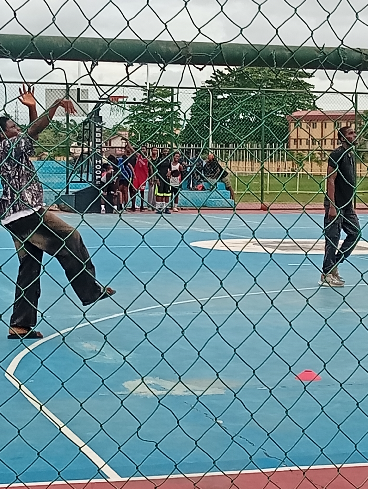A daytime, eye-level outdoor shot taken from behind a green metal wire fence depicts a group of people on a blue basketball court, in Ijanikin, Nigeria. On the left of the frame, a man wearing a patterned, long-sleeved shirt and dark pants appears to be mid-motion, his left arm extended upwards. In the background, a group of people are clustered near a blue bench, observing the activity. To the right of the group, another man wearing a black shirt and dark pants is walking along the court. An orange cone sits on the court. A basketball hoop and the word "HOOPS" is visible in the background. Buildings and trees are visible in the background, under a cloudy sky.