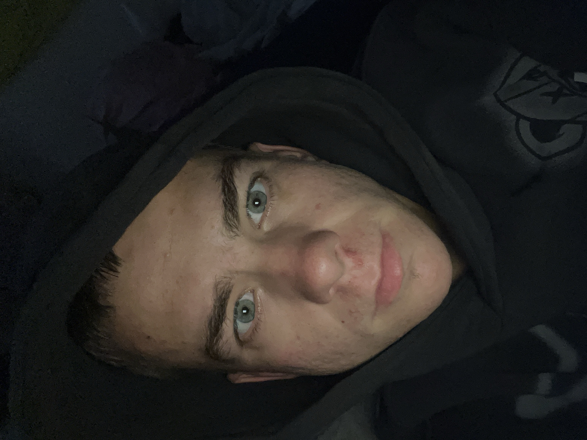 This is a close-up, self-portrait photograph of a young Caucasian male with light skin and short brown hair. He is wearing a dark hooded sweatshirt, the hood of which is pulled up around his head, obscuring the background. His eyes are a striking light blue-green and he is looking directly at the camera. His face shows some signs of acne, particularly on his cheeks and around his nose and mouth. The lighting is dim, creating a dark environment with harsh shadows, suggesting the photo was taken indoors or at night. The overall impression is casual and intimate. This photograph was taken in Velykyi Bychkiv, Ukraine.