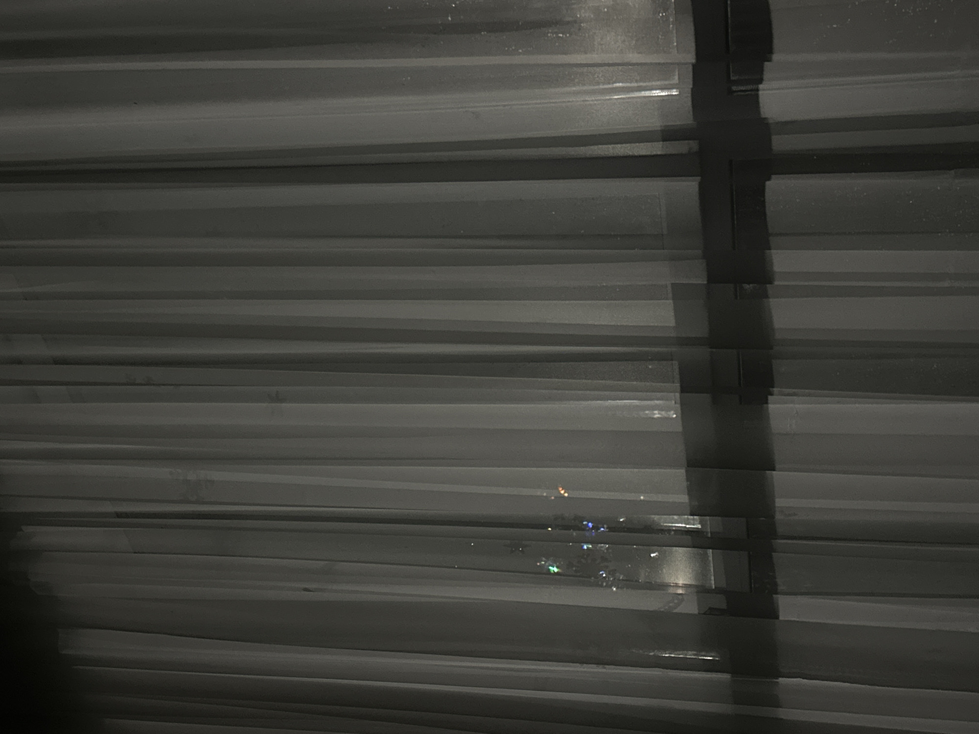 The image shows horizontal blinds in a dark environment. Light reflections are visible through the blinds, indicating an external light source. The blinds are partially closed. The scene appears to be indoors, with the blinds obscuring the view outside. The precise location is indeterminable without further context or visible landmarks.