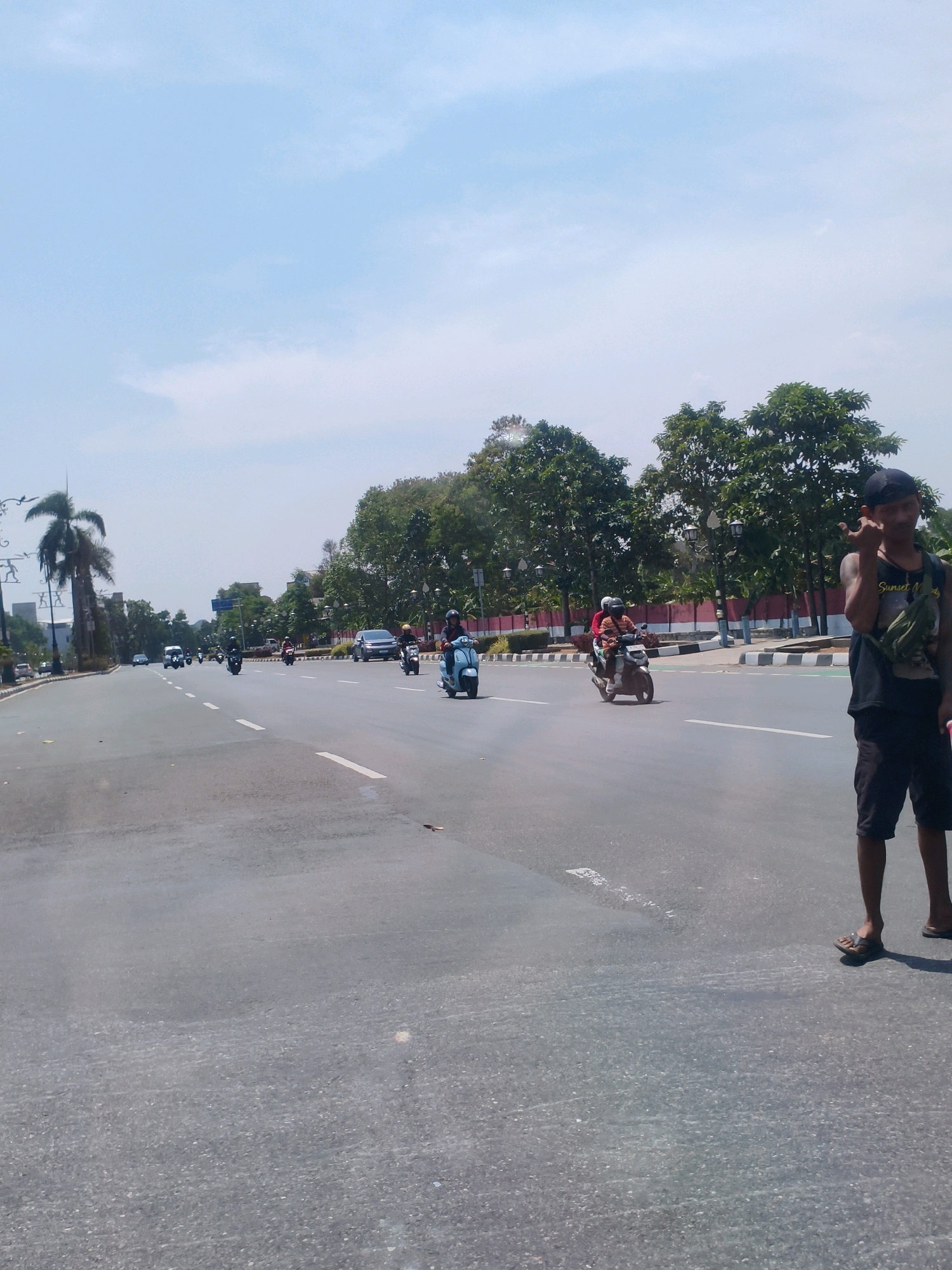 The image captures a street scene in Babakan Madang, Indonesia, under a clear blue sky with scattered clouds. The foreground features a paved road with white lane markings. On the right side of the road, a young man wearing dark shorts, a dark tank top with "Sunset" printed on it, a black baseball cap, and a green sling bag is standing. He is making a hand gesture with his right hand, showing his index and middle fingers extended. He is wearing flip-flops.
In the middle ground, several motorcycles are in motion on the road. One blue scooter is in the center lane, ridden by a person wearing a helmet. To the left of it, another motorcycle with two occupants, one of whom is wearing a helmet, is visible. Further down the road, more motorcycles and a car are traveling in the same direction.
The background of the image is lined with trees, some of which are palm trees. A red-and-white striped barrier runs along the side of the road, separating it from a pedestrian walkway or median. Streetlights are also visible along the roadside. The overall impression is a sunny day in a developing urban area, with moderate traffic on the main road.