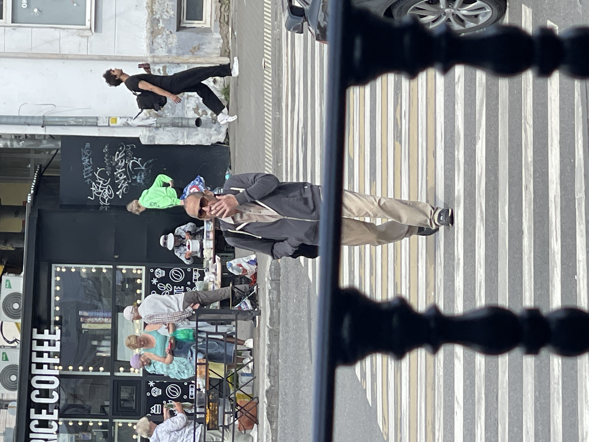 The image shows a street scene in Izhevsk, Russia, with a focus on a pedestrian crossing and the area around a cafe called "PRICE COFFEE." In the foreground, a man with sunglasses and a light-colored shirt under a gray hooded jacket is walking across the zebra crossing, smoking a cigarette. He is wearing light brown pants and dark shoes. Behind him, on the sidewalk, a young man with curly hair, wearing a black t-shirt and black pants, with a black backpack, is walking away from the camera, holding a can. Further back, several people are visible near the "PRICE COFFEE" establishment. A woman in a light blue top and a purple headscarf is interacting with another woman in a white shirt. Other individuals are standing or sitting at tables. The cafe has a black facade with "PRICE COFFEE" written in white letters. The background shows a typical urban environment with buildings and street infrastructure. The overall impression is of a casual day in a city.