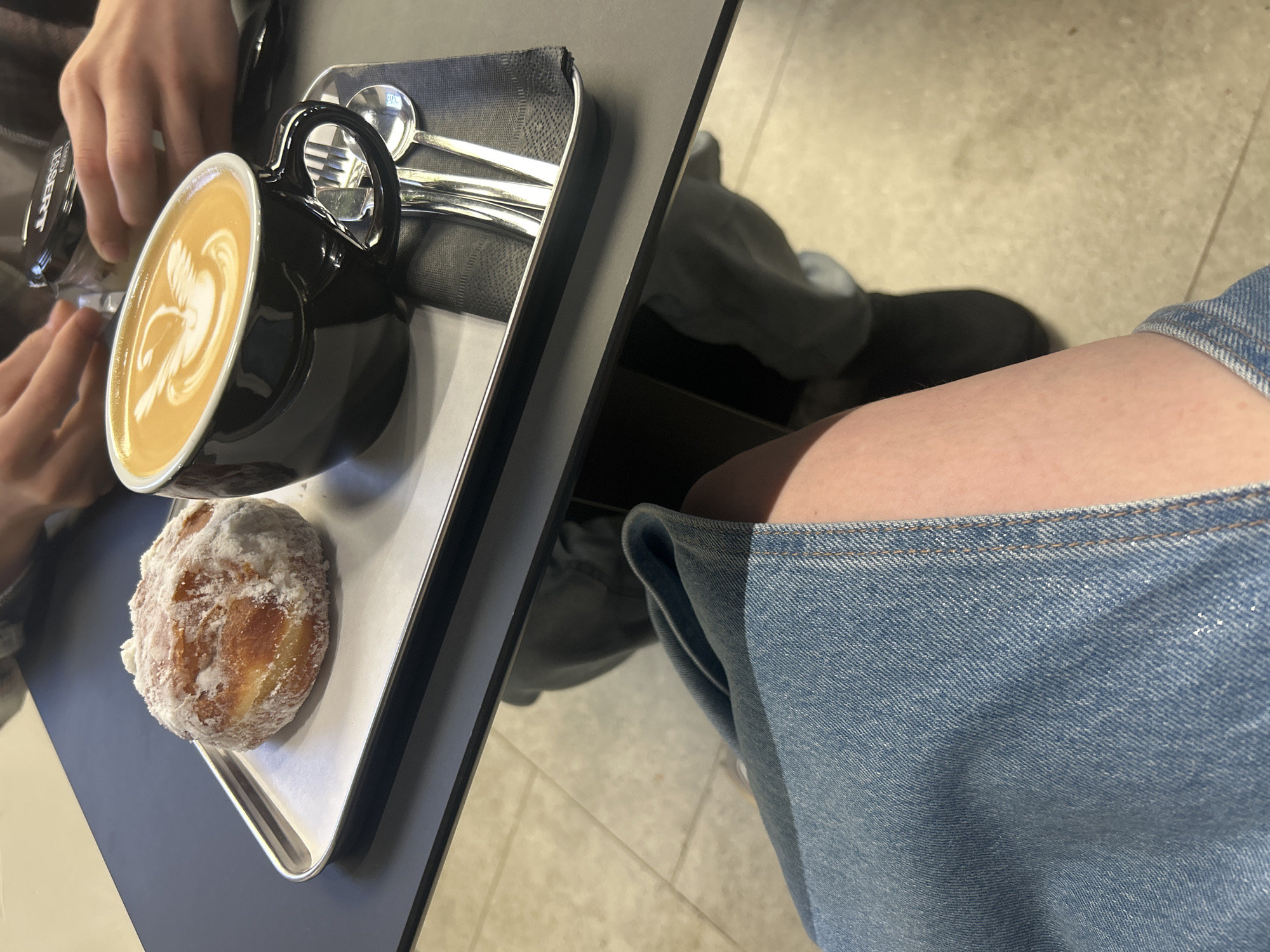 An overhead, medium shot shows a table with a tray of food and drinks. On the left, a sugary pastry sits on a metal tray. To the right of the pastry, a black coffee mug filled with latte art rests on a dark napkin. A fork and spoon are placed next to the mug. In the background, two individuals are visible, with one person's hands reaching towards a small dessert container. The foreground of the image shows a portion of a person's legs clad in denim jeans. The location is a cafe in Izhevsk, Russia.