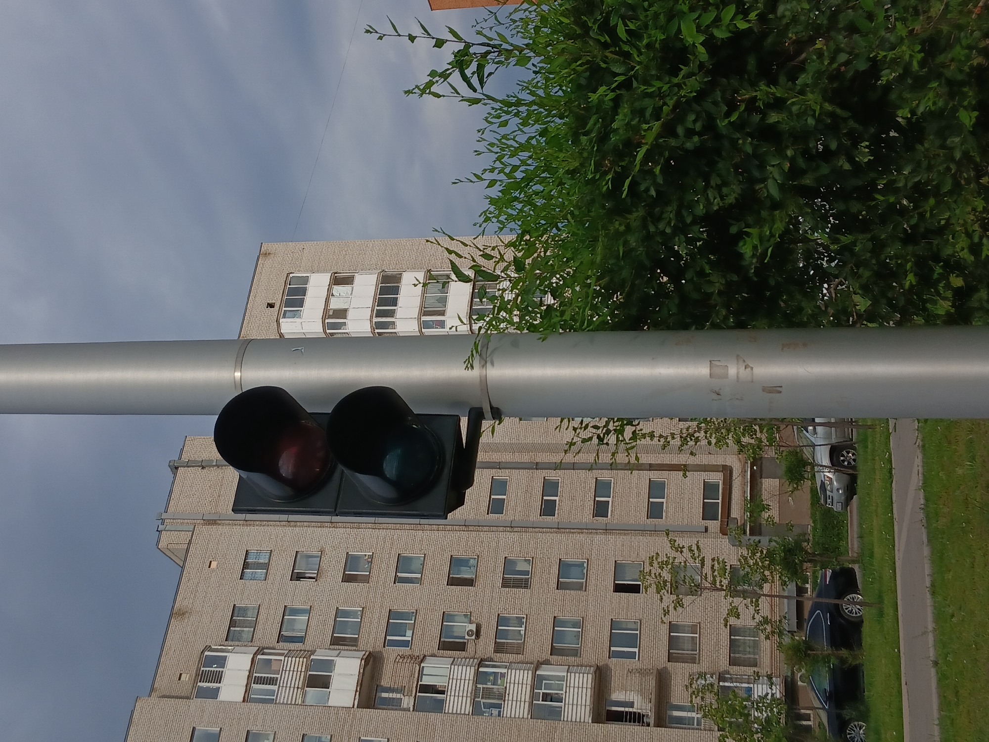 The image depicts a street scene with a traffic light and a multi-story apartment building in Astana, Kazakhstan. The traffic light, positioned on a silver pole, shows a red light illuminated at the top and a dark, unlit green light below. The apartment building is constructed with light-colored brick and features multiple floors with numerous windows and balconies. In the foreground, a dense green hedge is visible on the right side of the frame. On the left side of the frame, a black car and a silver SUV are parked along the side of the road. The sky is overcast with a mix of gray and white clouds. The area appears to be a residential or urban environment.