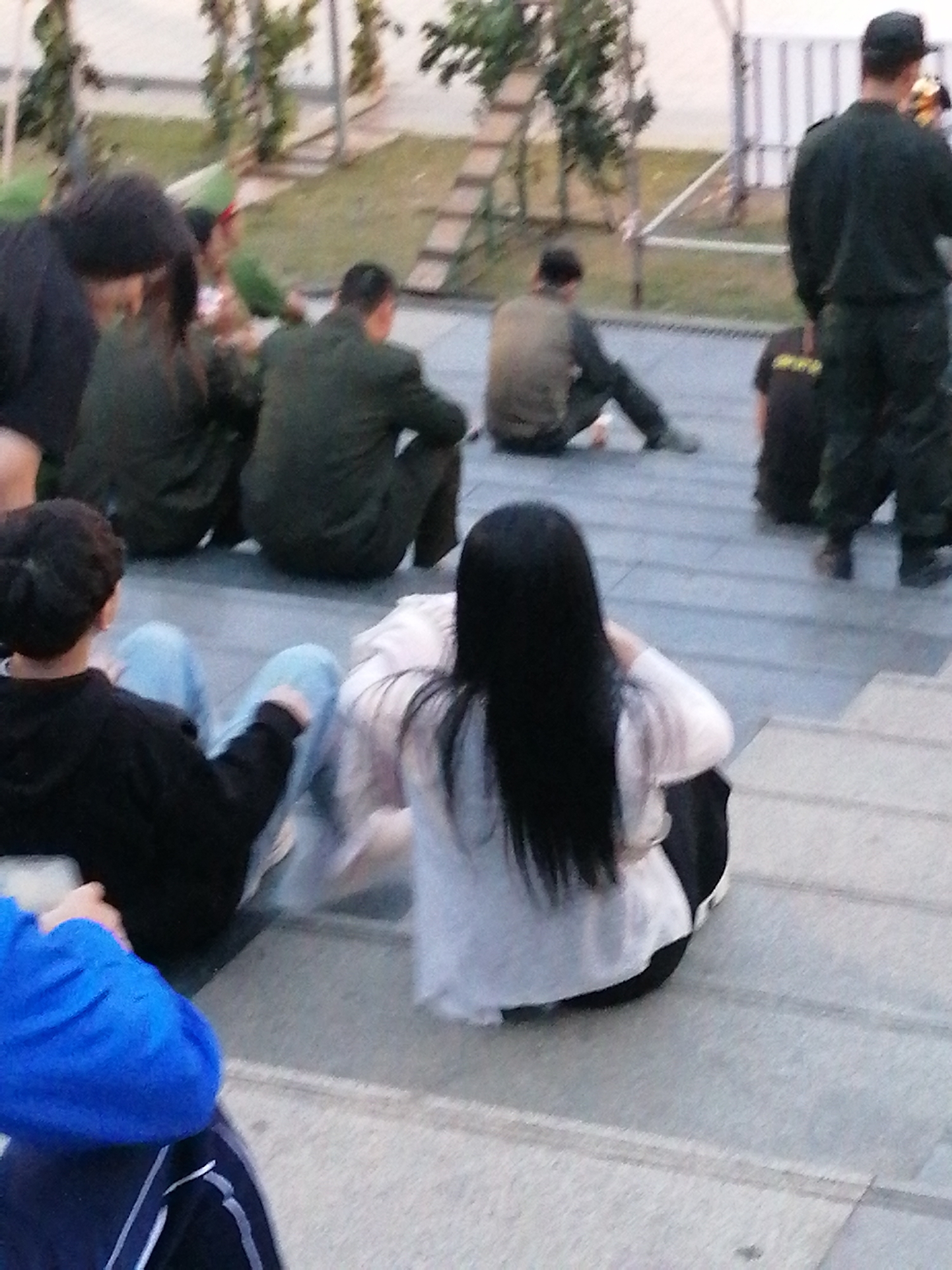A group of people are sitting on outdoor steps, some facing away from the camera, others in profile. The scene appears to be a public area, possibly a park or a plaza, with some greenery and a ladder-like structure in the background. The lighting suggests it is either early morning or late afternoon.
In the foreground, a person wearing a blue jacket and dark pants sits with their legs spread. To their right, a person with long dark hair, wearing a white top and dark pants, is also seated. Further to the right, partially obscured, are two individuals in dark green uniforms, sitting on the steps. One of them is facing away from the camera.
In the middle ground, a person in a light brown jacket is seated on the steps, facing to the left. To their right, another person in a dark uniform is standing, facing away from the camera.
The overall atmosphere is casual, with people appearing to be resting or waiting. There are no obvious indications of a specific event, but the presence of uniformed individuals might suggest a security or organizational role. The location context of Da Lat, Vietnam, does not offer specific visual cues within the image itself. No visible text is present in the image.
