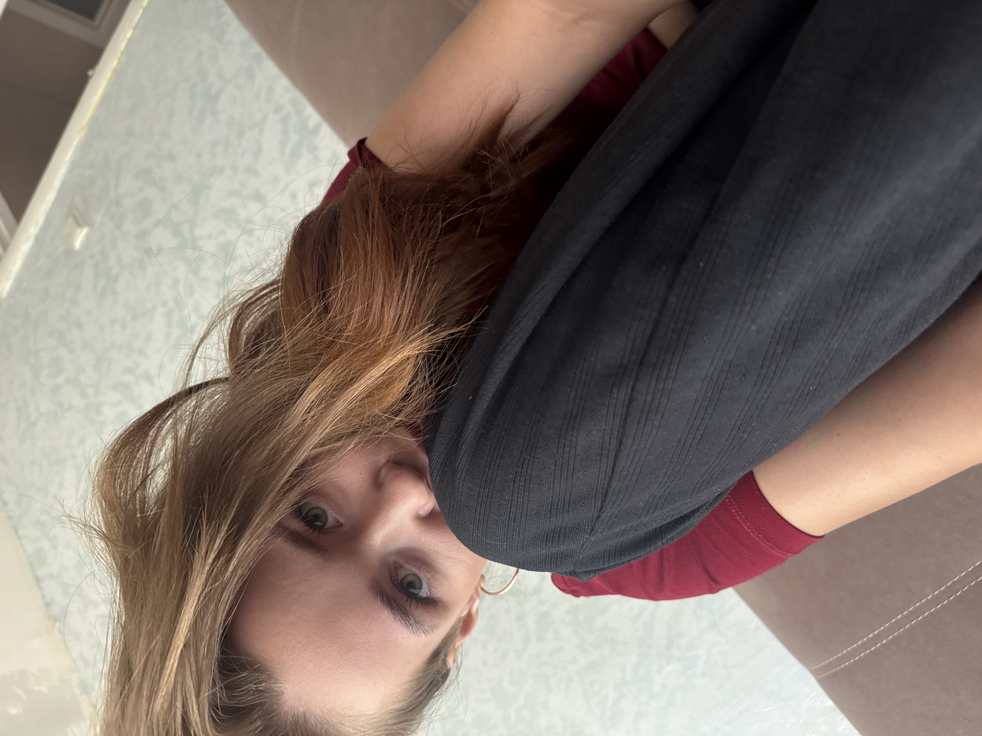 A close-up, rotated shot focuses on the face of a young woman with shoulder-length, auburn hair. Her eyes, a striking green, are looking directly at the camera. She appears to be resting her head on her arm, with her hair falling around her face. She is wearing a dark-colored, ribbed long-sleeved shirt over a maroon t-shirt. A large hoop earring is visible in her right ear. The background consists of a light-colored, textured wall with a subtle pattern, and a hint of a brown upholstered surface, possibly a couch, is visible on the right. The lighting suggests an indoor setting, likely during daylight hours. The woman's expression is calm and direct. There is no visible text in the image. The overall impression is a casual, personal snapshot.