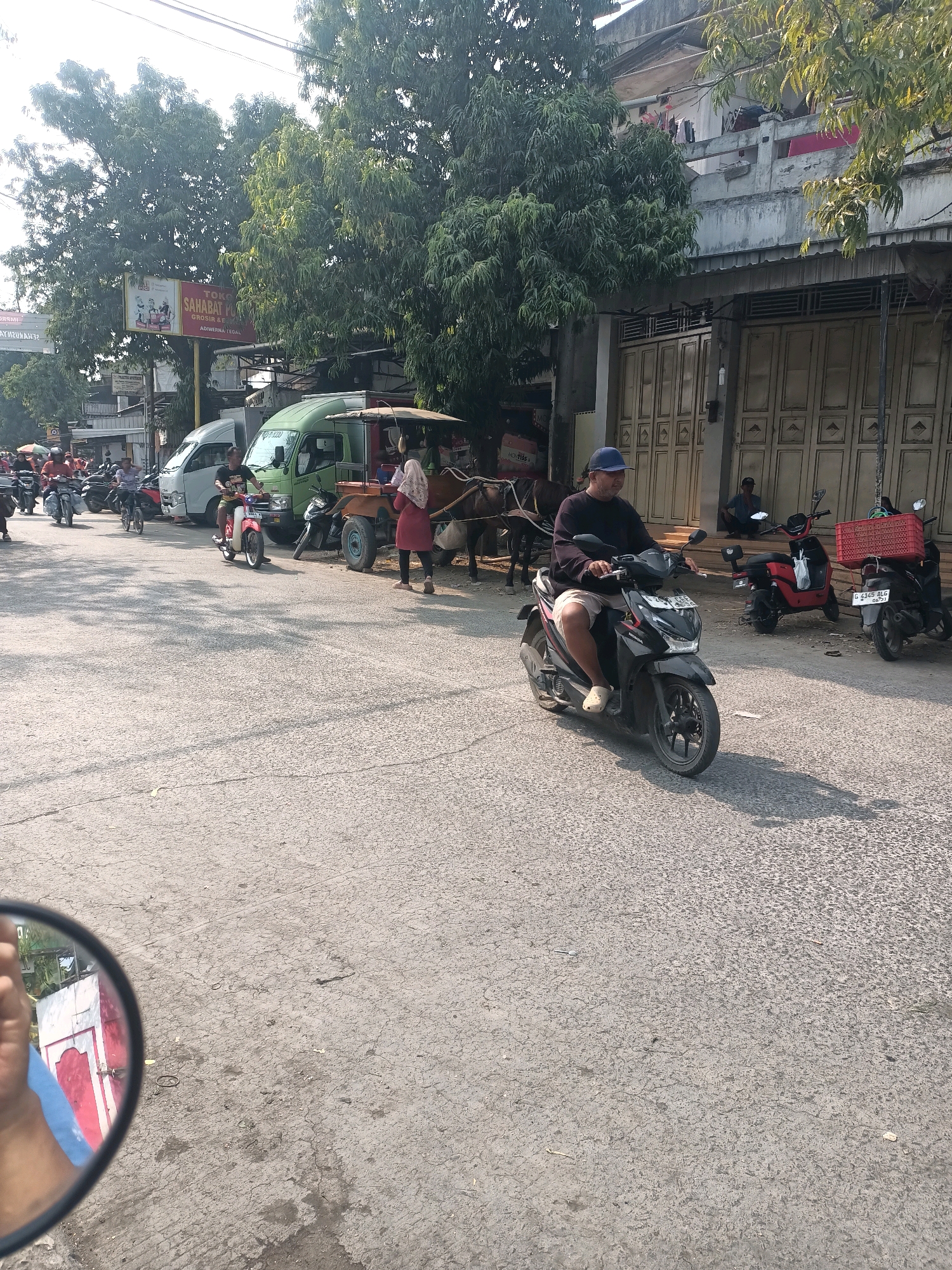 The image captures a street scene in Ujungrusi, Indonesia, under bright sunlight casting long shadows. A man wearing a blue cap and light-colored shorts rides a black motorcycle in the foreground. To his right, a red motorcycle is parked, and further back, another motorcycle with a red crate attached to its rear is visible. Behind the parked motorcycles, a dark-haired individual sits on a low wall. In the mid-ground, a green truck is parked, and beside it, a horse-drawn cart is positioned. A woman in a pink hijab and a red tunic stands near the cart, holding onto one of the horses. Several other motorcycles and a white van are also present on the street. Trees line the left side of the road, and buildings with closed doors and windows are situated on the right. A red and white structure is reflected in the side mirror of a vehicle, suggesting the perspective is from a moving vehicle. A banner with Indonesian text and images is attached to a pole amidst the trees. The road surface is asphalt, showing signs of wear and tear.