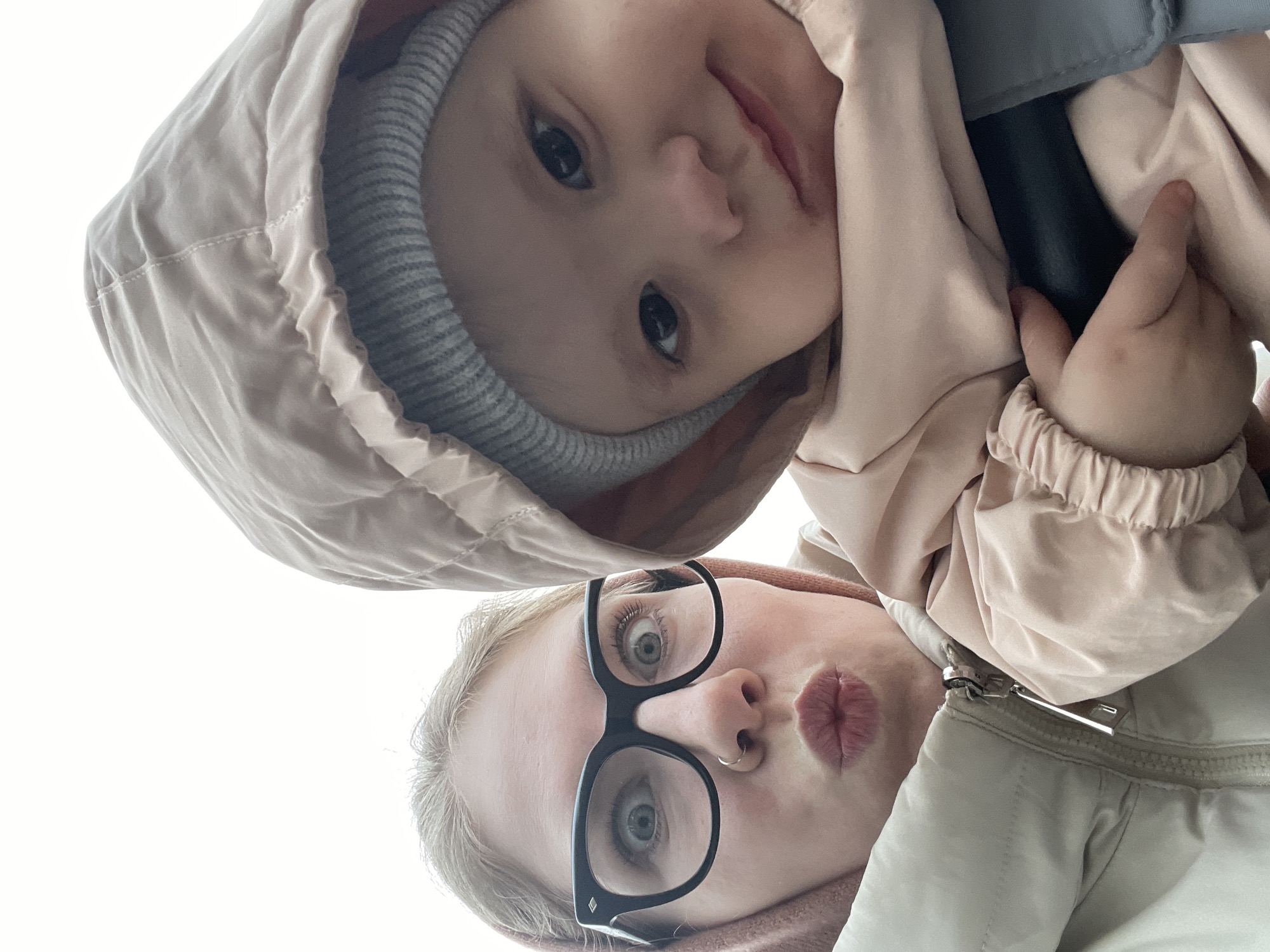 Two individuals, an adult and a child, are positioned closely together, with the camera angle directed upwards towards them. The child, wearing a light pink hooded jacket and a gray beanie, is situated above the adult. The adult is wearing glasses and a light-colored jacket. The background is a bright, overcast sky. There is no evidence to suggest that this image was taken in Izhevsk, Russia.