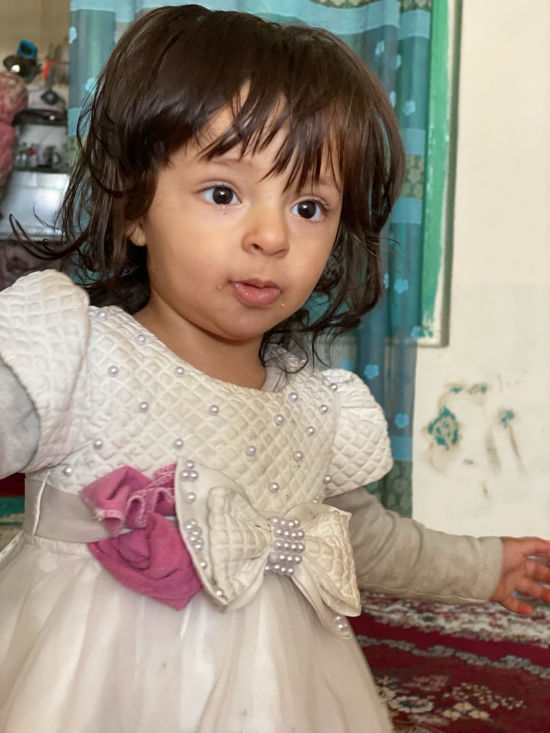 A young child is the focus of the image, wearing a light-colored dress adorned with pearls and a large bow. The dress also features long sleeves. The child has dark hair and appears to be indoors. The background includes a curtain with a patterned design and a wall that appears to be aged. There is also a carpet visible in the lower part of the image. It is not possible to determine the location in Kandahar, Afghanistan, from the information provided.