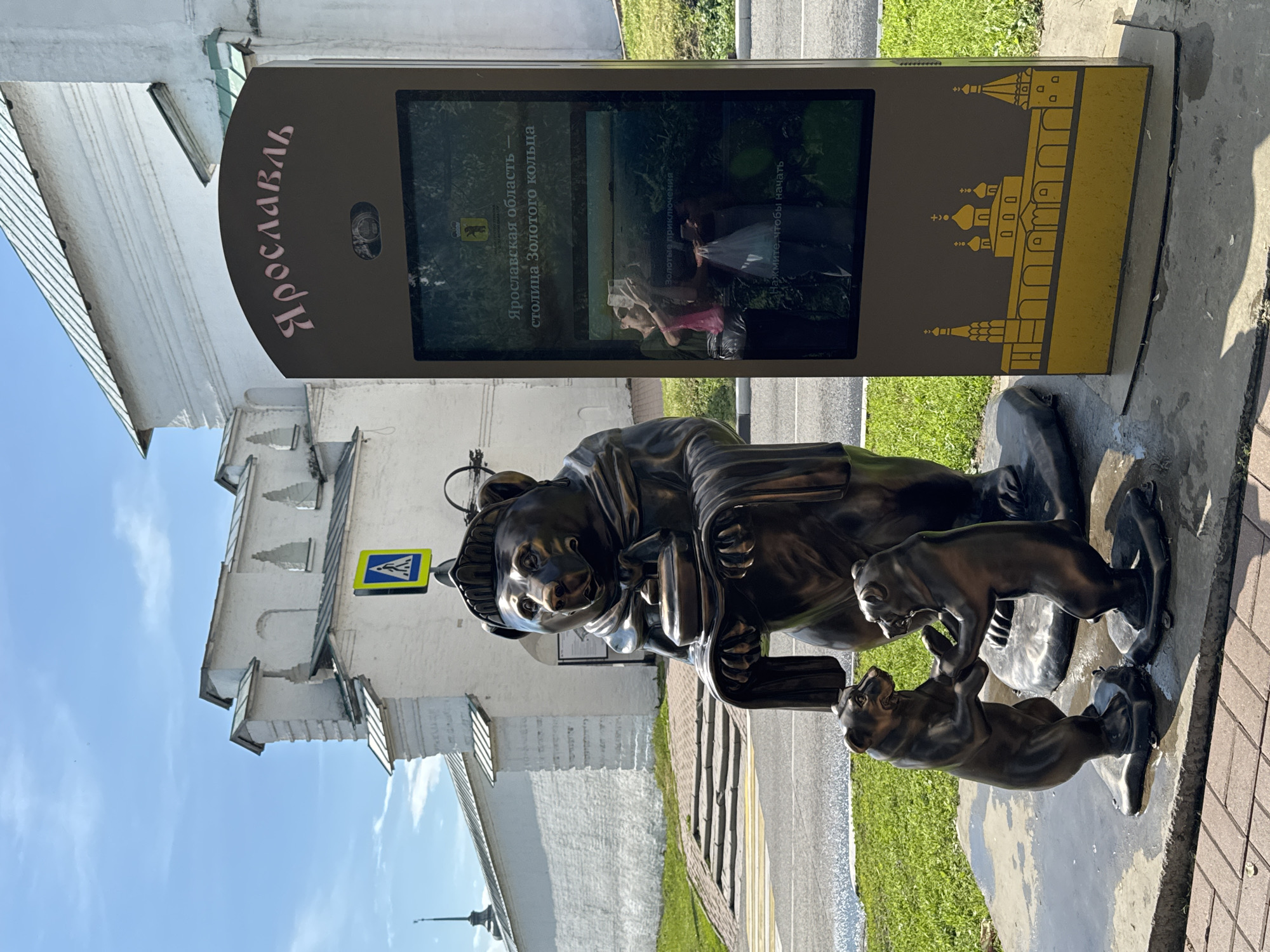In Rusanovo, Russia, a bronze sculpture of a mother bear holding a small pot is depicted with two smaller bear cubs dancing at her feet. The sculpture is located outdoors on a concrete pedestal, with a paved area and grass surrounding it. Behind the sculpture is a digital kiosk displaying information about Yaroslavl Oblast, the capital of the Golden Ring. The kiosk features an image of the Yaroslavl coat of arms and text in Russian. In the background, a white building with a green roof and arched windows is visible, along with a pedestrian crossing sign. The sky is blue with some clouds.