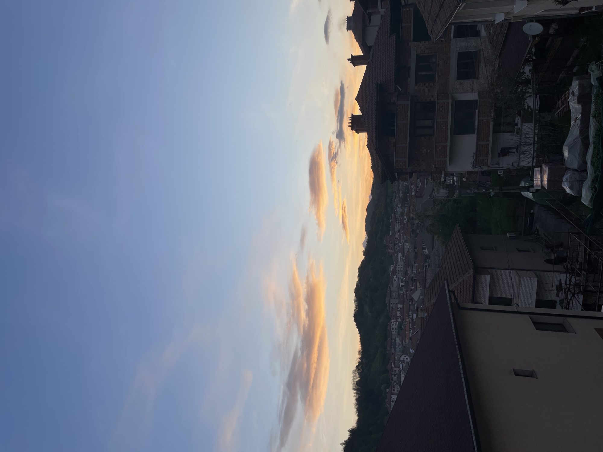 The image presents a landscape view, likely taken at sunset in Zlatograd, Bulgaria. The sky, occupying a large portion of the left side, transitions from a clear, light blue to hues of orange and peach near the horizon, where wispy clouds are illuminated by the setting sun.
On the right side, the image is dominated by a dense collection of buildings and houses, characteristic of a European town. Red-tiled roofs are visible, indicative of traditional architecture. The buildings ascend a hill, creating a terraced effect. Some structures appear to be under construction, featuring exposed brickwork. Green foliage, likely trees and vegetation, is interspersed among the buildings.
The foreground includes the top of a building with a dark roof, partially obscuring the view of the town below. The image is oriented vertically, with the sky on the left and the urban landscape on the right.