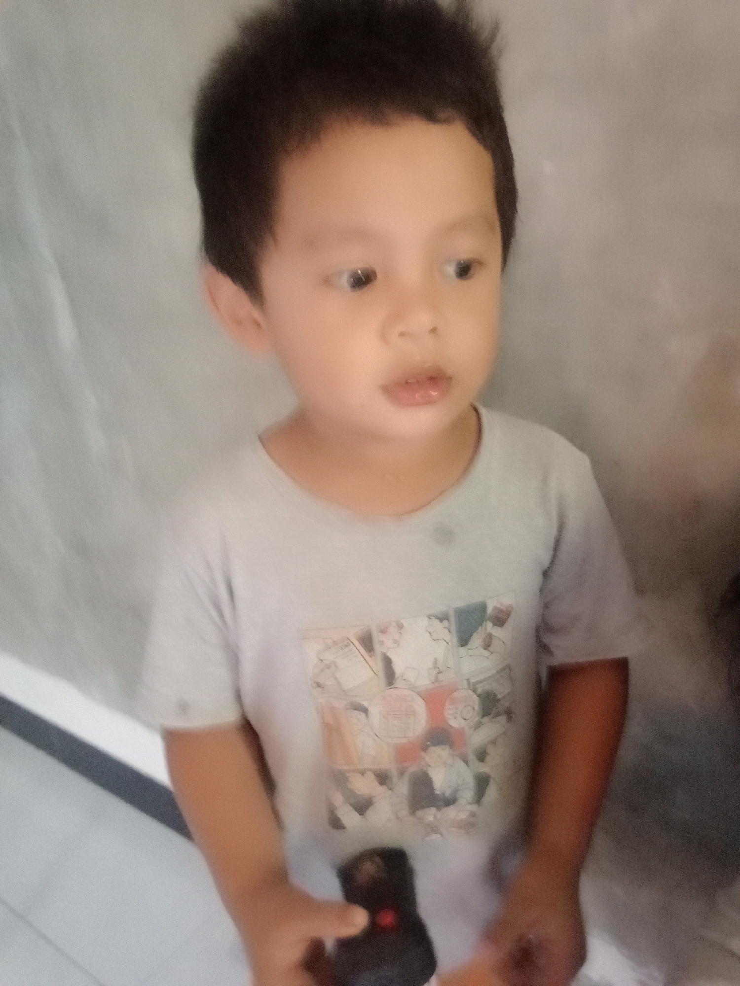 A young boy with dark hair is pictured from the chest up, facing slightly to the right of the camera. He is wearing a light gray t-shirt with a graphic print of what appears to be comic book panels. His mouth is slightly open, and his eyes are looking to his right. His right hand is visible, holding a small black object with a red light, possibly a remote control. The background is a textured, light gray wall, with a draped white fabric on the left side of the frame. The lighting is soft and diffused. The image appears to have been taken indoors. There is no information to indicate the location in Muara, Indonesia.