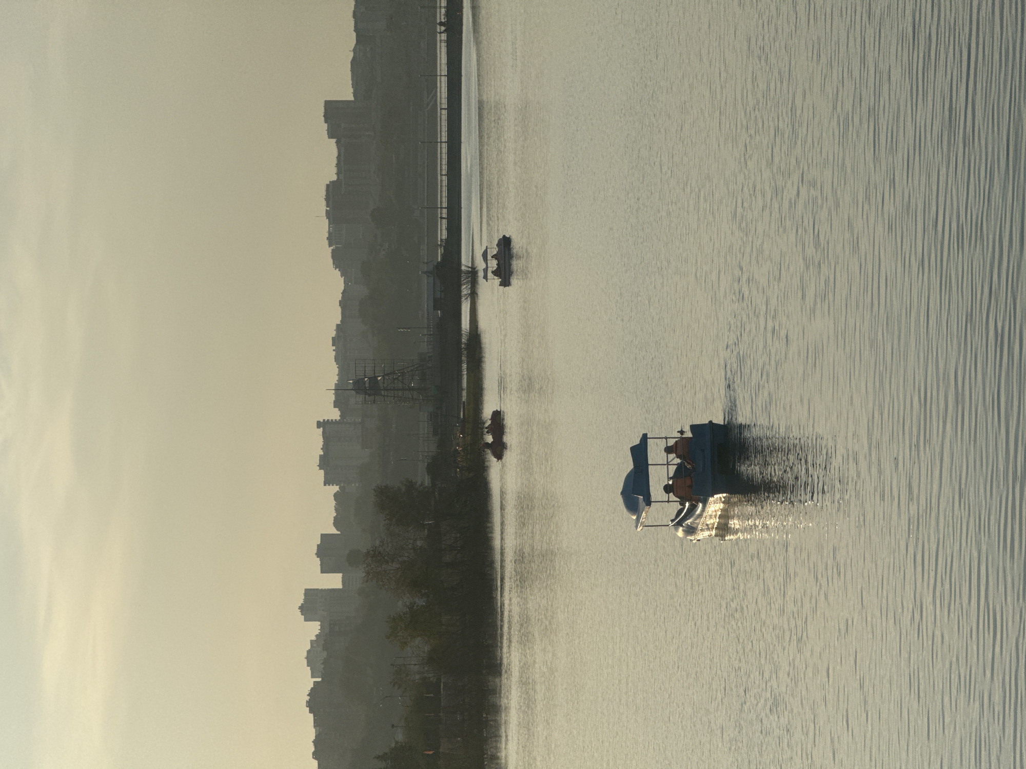 The image captures a serene view of the Angara River in Irkutsk, Russia, during the golden hour. In the foreground, a blue pedal boat with a canopy is navigating the calm waters. Two individuals, wearing orange life vests, are seated inside, presumably pedaling the boat. The water's surface is gently rippled, reflecting the soft, hazy sunlight that bathes the scene.
Further in the distance, another pedal boat with two occupants is visible, also enjoying a leisurely ride on the river. Beyond the water, the cityscape of Irkutsk unfolds. A line of buildings, ranging from apartment complexes to possibly older structures, forms the skyline. Trees and vegetation line the riverbank, creating a verdant transition between the water and the urban environment. The overall atmosphere is peaceful and tranquil, with the muted colors and soft light suggesting either early morning or late evening. A bridge can be seen spanning the river in the mid-ground, with streetlights lining its length. The sky is a pale, washed-out yellow, indicating either a misty or overcast condition.