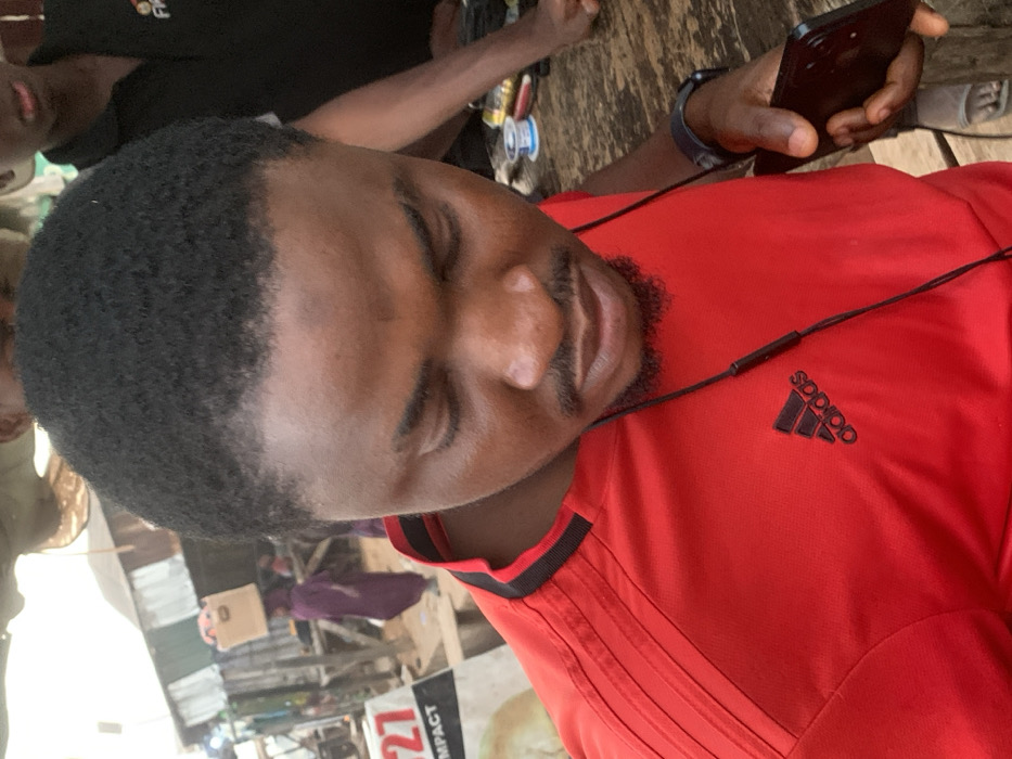 A man is angled, looking down at a black phone in his right hand. He's wearing a red Adidas shirt with black stripes and the Adidas logo. Black earphone wires are visible across his chest. His eyes are closed, and he has short dark hair and a beard. A wrist-worn device is visible on his left arm. People and structures form a busy outdoor background.