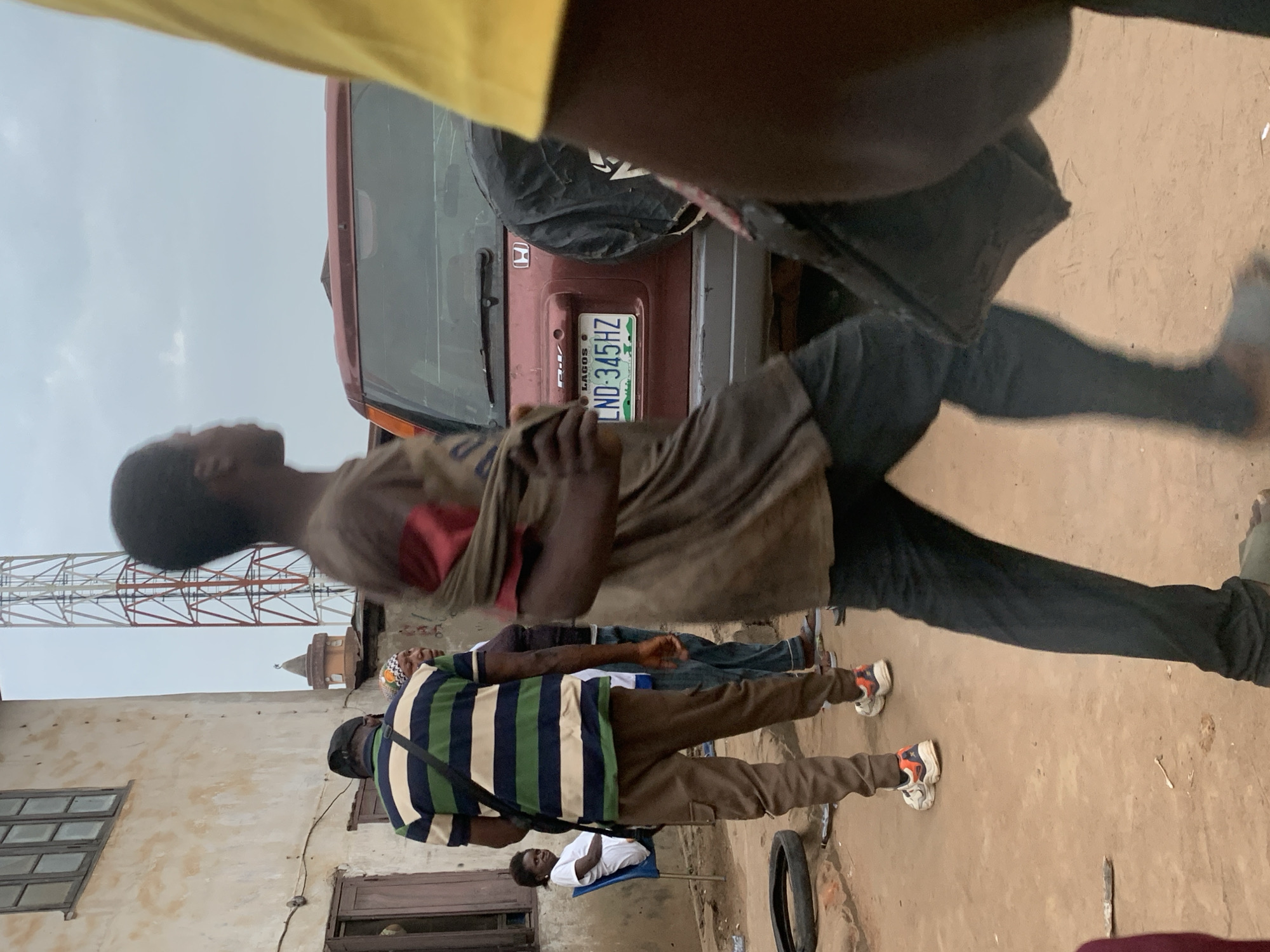 The image depicts an outdoor scene in Shagamu, Nigeria, likely a street or an open area adjacent to buildings. In the foreground, a young man is walking away from the camera, wearing a dirtied t-shirt with a red cloth draped over his left shoulder and dark pants. He is carrying a black bag. To his left, another person is partially visible in a yellow shirt.
In the middle ground, a red SUV is parked, its rear visible. A license plate reads "LAGOS LND 345 HZ." To the left of the SUV, a man wearing a striped polo shirt (green, blue, and white) and brown pants walks away from the camera, carrying a black bag over his shoulder. Beside him, a woman in a headscarf and another person stand. Further left, a woman sits on a blue chair, wearing a white shirt.
The environment appears somewhat unkempt, with a tire lying on the ground to the left of the man in the striped shirt. Buildings with weathered walls and windows are visible in the background, along with a tall transmission tower. The sky is overcast, suggesting a cloudy day. The overall impression is of a candid street scene in a Nigerian city.