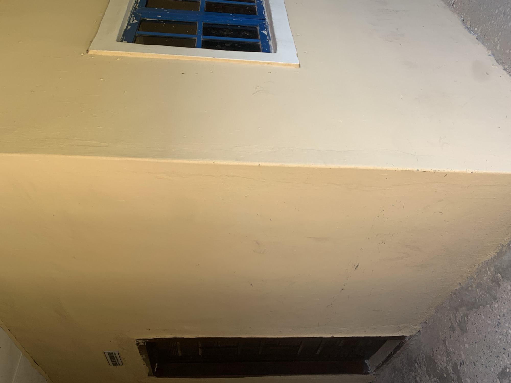 The image depicts a corner of a building with cream-colored walls. To the left, a dark wooden door is visible, set into a recessed doorway. Above the door, a small rectangular sign with text is affixed to the wall. To the right, a window with blue-painted panes and a white frame is present. The floor in the foreground is a concrete surface with a speckled pattern. The scene appears to be an exterior view of a building in Jalingo, Nigeria, likely a public or commercial structure given the presence of the sign and window. The lighting suggests it is daytime.