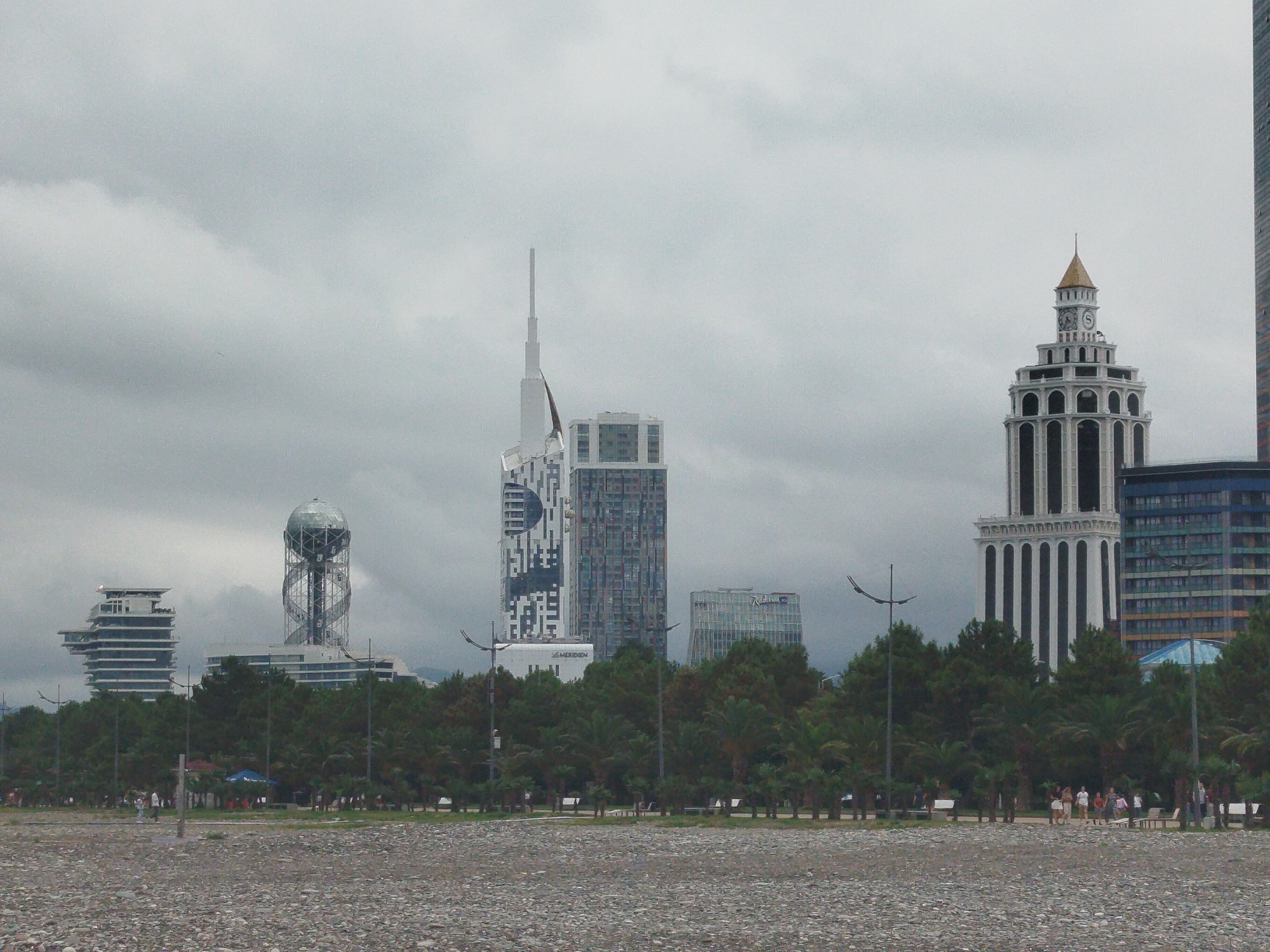 The image depicts a scene in Batumi, Georgia, featuring a shoreline of pebbles in the foreground. Beyond the shore is a line of lush green trees and palm trees. Behind the trees, a cluster of modern skyscrapers rises against a cloudy, overcast sky. The buildings vary in architectural style and height, with notable structures including a tower resembling a DNA helix topped with a spherical observatory, a white building with a spire, and a distinctive tower with arched windows and a gold-capped roof. The middle ground shows a promenade or park area with lampposts and a few scattered individuals walking or standing. The overall atmosphere is subdued due to the cloudy weather.