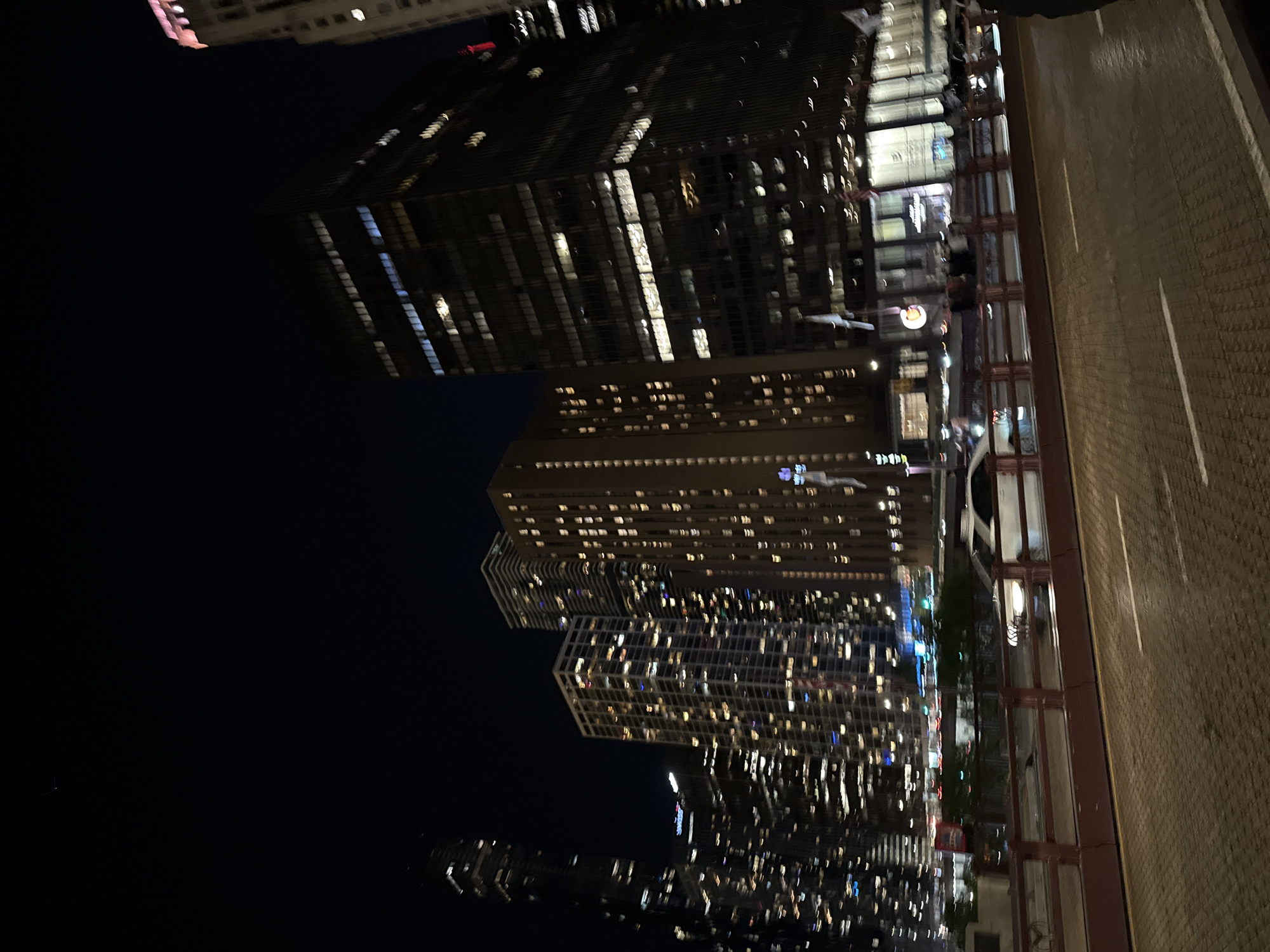At night, the image depicts an upward-looking cityscape from what appears to be a walkway, possibly near the river. Buildings of varying heights, all with many windows illuminated, dominate the scene, giving off a bright glow against the dark sky. The walkway itself is visible, with stone patterns and a guardrail on the right.