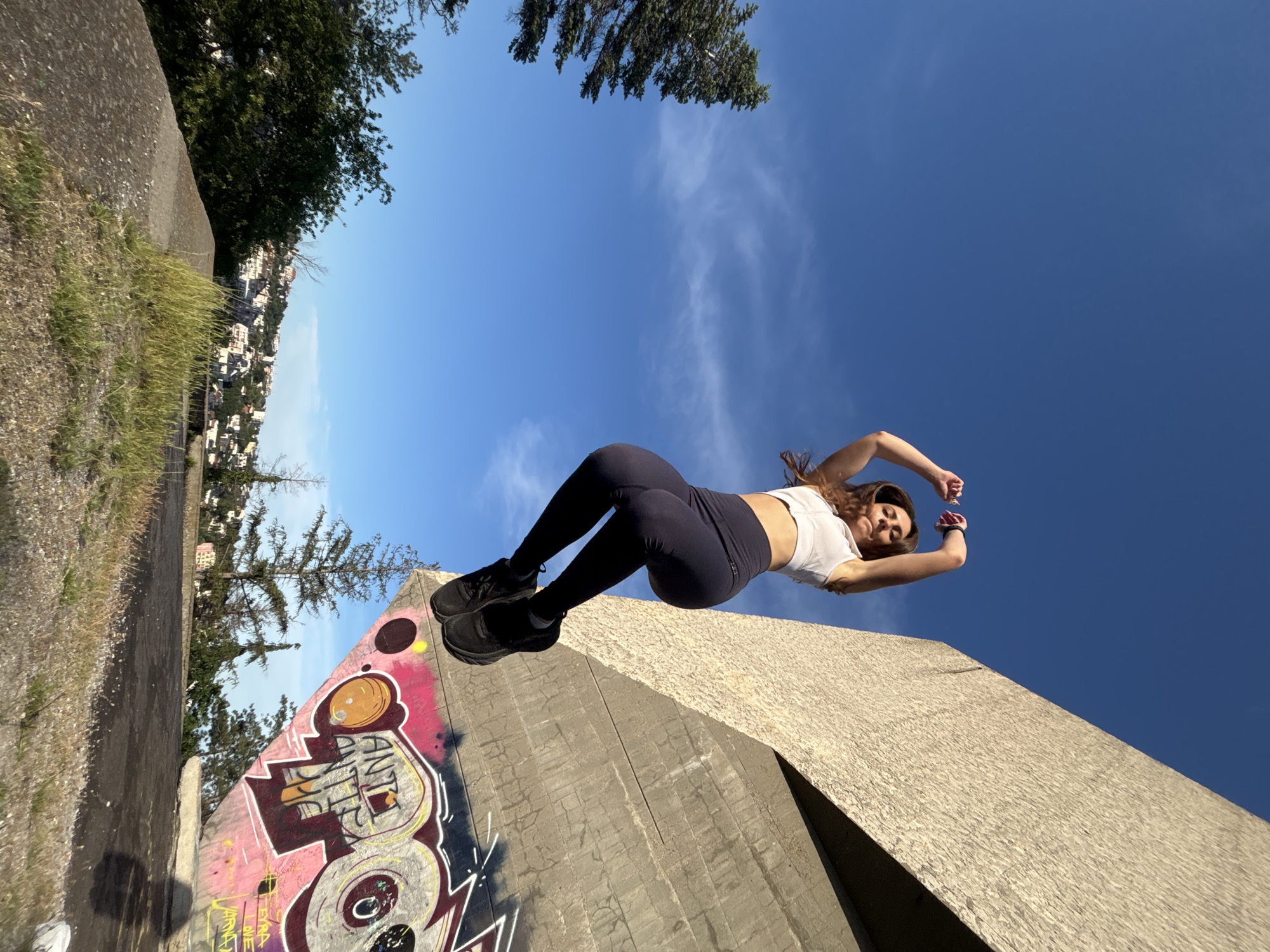 A young woman is captured mid-air, performing a jump against a clear blue sky with scattered clouds. She is wearing a white crop top and dark leggings, with black sneakers. Her arms are raised above her head, and her legs are bent. The background features a large, angular concrete structure with graffiti on its surface. To the left of the structure, evergreen trees and a distant cityscape are visible, with buildings dotting a hillside. The overall setting appears to be an outdoor urban environment. The graffiti on the concrete structure includes the words "ANTI ANTI" and other stylized lettering in various colors, including pink, yellow, and white. The city in the background is identifiable as Varna, Bulgaria.