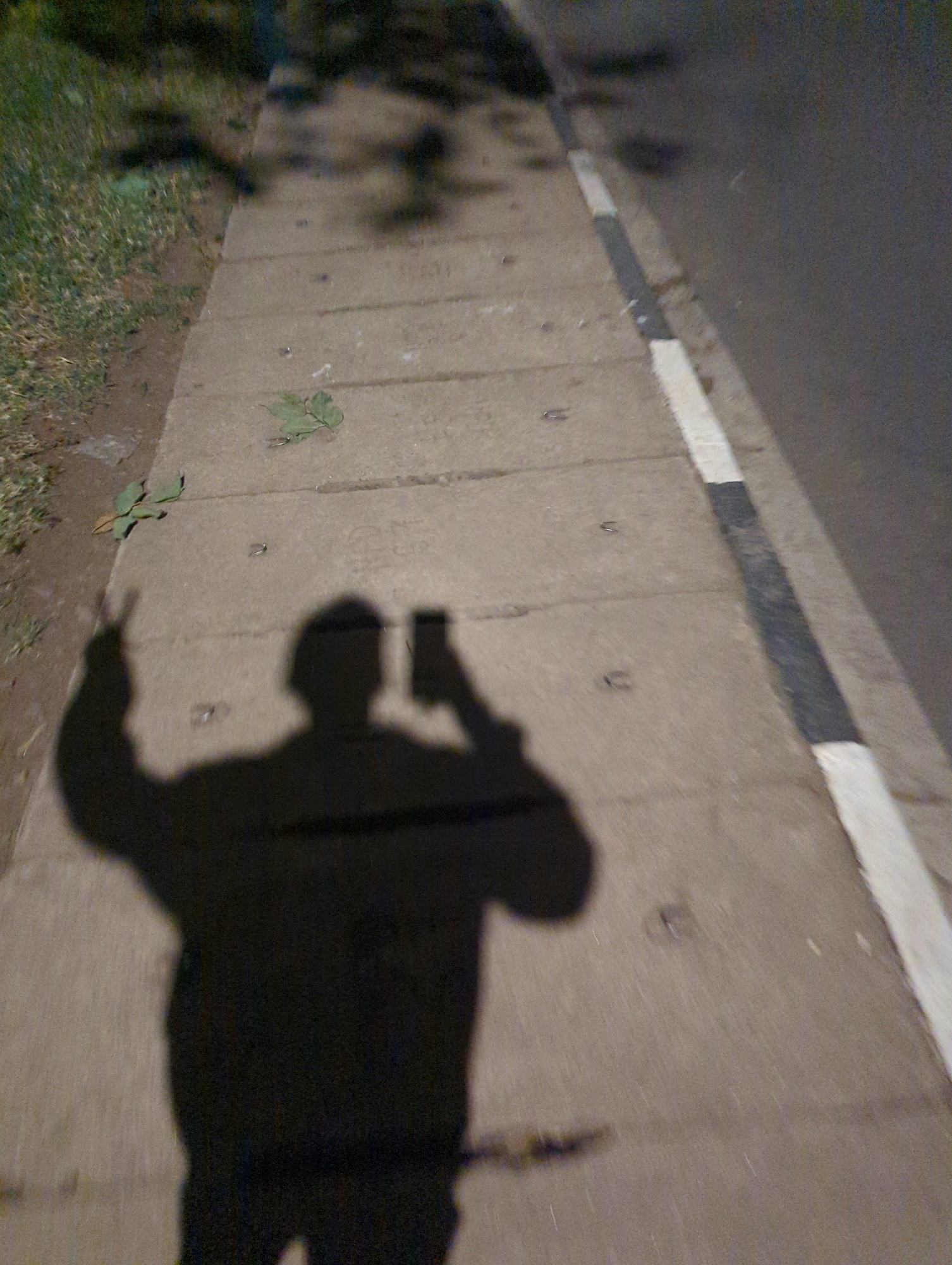 An undated, low-resolution, dimly lit, ground-level outdoor photograph captures the shadow of a person taking a selfie while standing on a concrete sidewalk. The sidewalk is composed of rectangular slabs, and a few dried leaves are visible on its surface. To the left of the sidewalk, a patch of grass and dirt is visible. To the right, the sidewalk meets a paved road with a distinct white and black painted curb. The person's shadow is cast in silhouette, showing them holding a smartphone in their right hand and raising their left hand with two fingers extended in a peace sign. The shadow extends across the sidewalk and onto the edge of the road. The ambient light suggests it is either dusk or nighttime, with a dappled shadow of tree foliage visible in the upper portion of the image. The scene appears to be a public walkway in Musanze District, Rwanda.