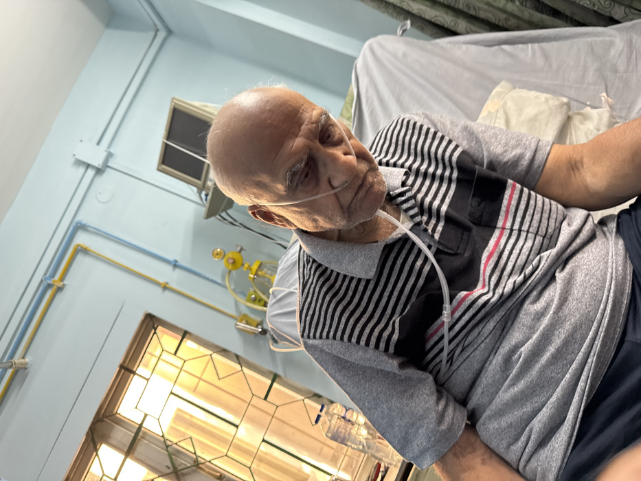 The image, taken with a tilted perspective, captures an elderly man in what appears to be a medical setting, possibly a hospital room or home care in Kolkata, India. The man, who is bald with white eyebrows, is seen in an upright or semi-reclined position, looking towards the right side of the frame with a somber or tired expression. He is wearing a nasal cannula, indicating he is receiving oxygen.
He is dressed in a short-sleeved polo shirt, which is predominantly grey with a distinctive pattern of black and white stripes across the chest and sleeves. His lower attire appears to be dark blue or black shorts or pants.
The background features light blue walls with exposed utility pipes—one blue and one yellow—running along them, commonly associated with medical gas lines. Behind the man's head, a yellow oxygen flowmeter and humidifier unit are visible, connected to these pipes. A window with a decorative metal grill allows bright natural light into the room, suggesting it is daytime. An air conditioner unit or vent is also visible on the wall. Next to the man, a clear plastic bottle, possibly containing water for humidification or an IV solution, is placed. In the upper right background, part of a bed or recliner with light-colored bedding and a patterned curtain can be seen. No discernible text is visible in the image.