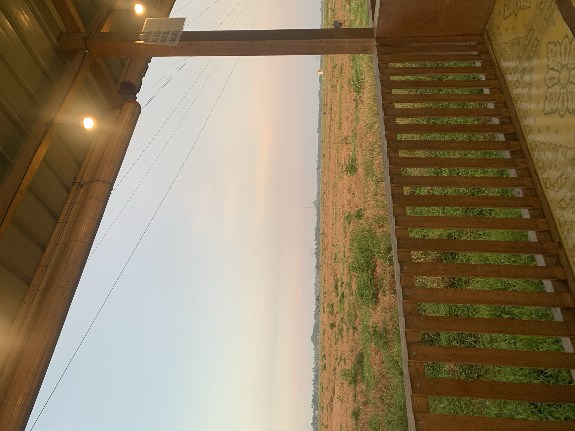 A wide shot captures a serene landscape at dusk, viewed from a rustic wooden veranda in Kualadua, Indonesia. The sky transitions from a pale blue to soft pinks and oranges, indicating the setting sun. A vast, dry field, dotted with patches of green vegetation, stretches to the horizon. In the distance, a line of trees and a few scattered buildings are visible. The veranda, constructed with natural wood, features a simple railing and a patterned yellow mat on the floor. Overhead, a metal roof is supported by wooden beams, and warm lights are illuminated. A sign with Indonesian text hangs from a wooden post on the right side of the frame. The overall scene conveys a peaceful, rural atmosphere.