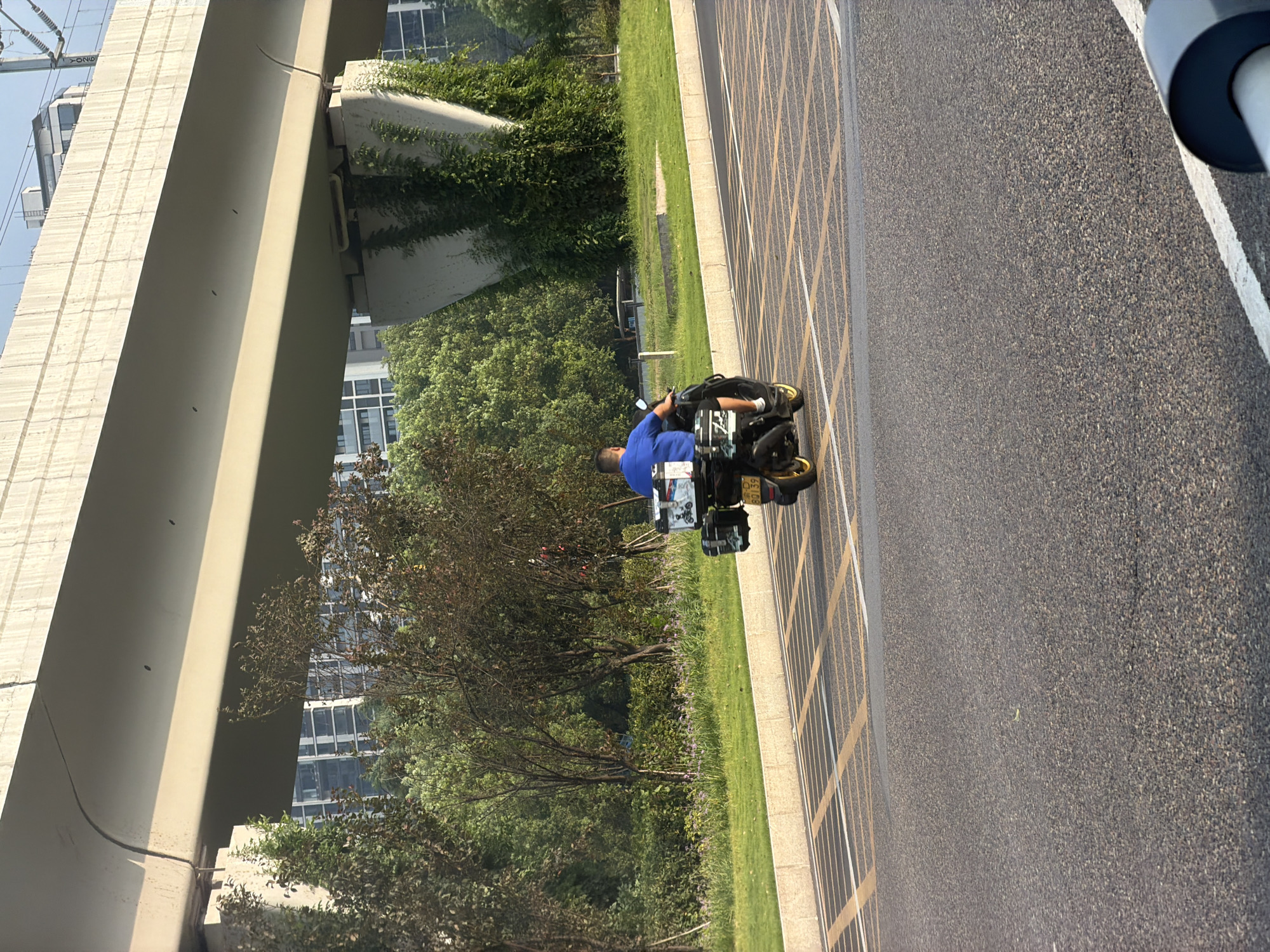 A man in a blue shirt rides a black scooter with two storage boxes mounted on the back. He is on a paved road with yellow and white directional markings. The road is bordered by a grassy area and a concrete barrier. In the background, there are trees, buildings, and an elevated concrete structure, possibly a bridge or overpass. The lighting suggests it is daytime. The license plate on the scooter reads "B0139".