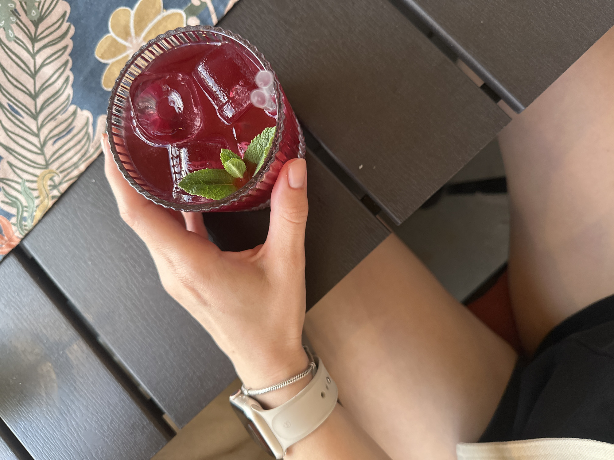 A person is holding a red beverage with ice cubes and mint leaves in a glass. The beverage appears to be on a dark gray slatted table outdoors. The person is wearing a white smartwatch and a thin silver bracelet. The person is wearing shorts with a white trim. In the background, there is a patterned tablecloth with floral and leaf designs. The location cannot be determined to be Perm, Russia from the image.