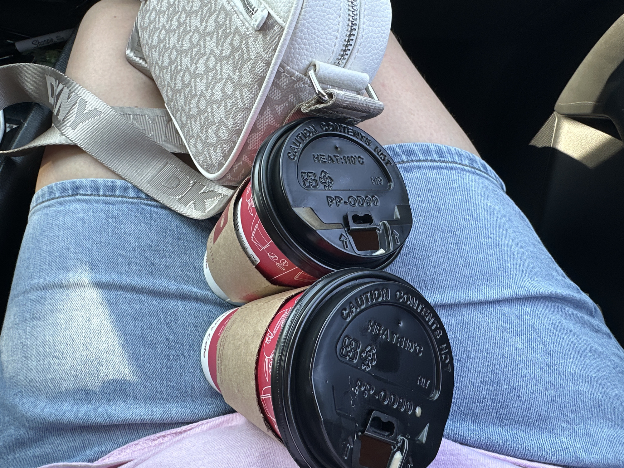 The image shows a close-up view of two disposable coffee cups with black lids resting on a person's lap, who is wearing blue denim jeans and a pink shirt. A light-colored purse with a textured pattern and a silver zipper is visible on the right side of the frame, connected by a beige strap with embossed lettering. The coffee cup lids have embossed warnings in English, including "CAUTION CONTENTS HOT" and "HEATING MIC." The lighting suggests an outdoor setting, possibly a car interior with sunlight casting shadows on the jeans. The location is identified as Lynnwood, United States of America.